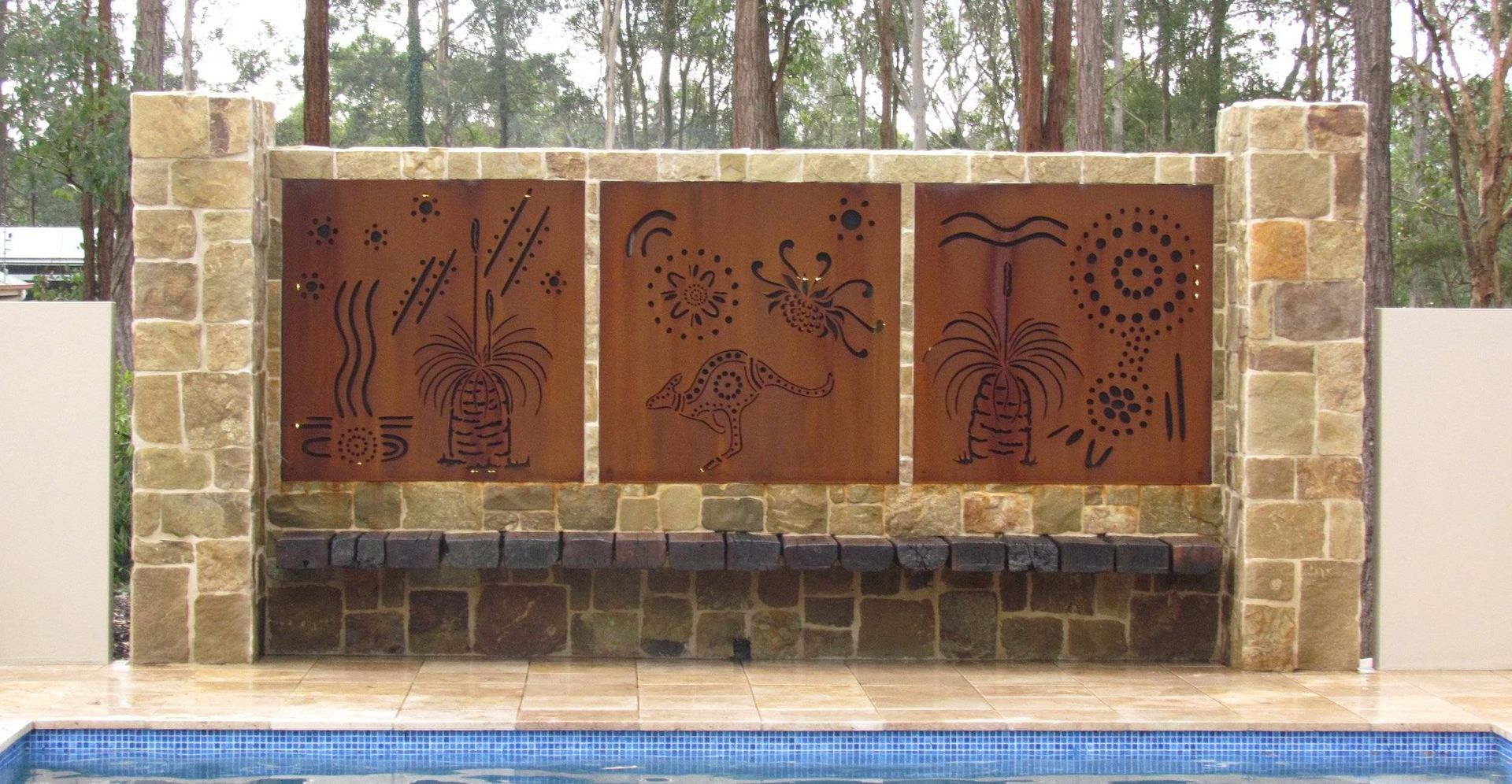 Three metal panels with cut-out designs set in a stone wall above a pool. Forest background.