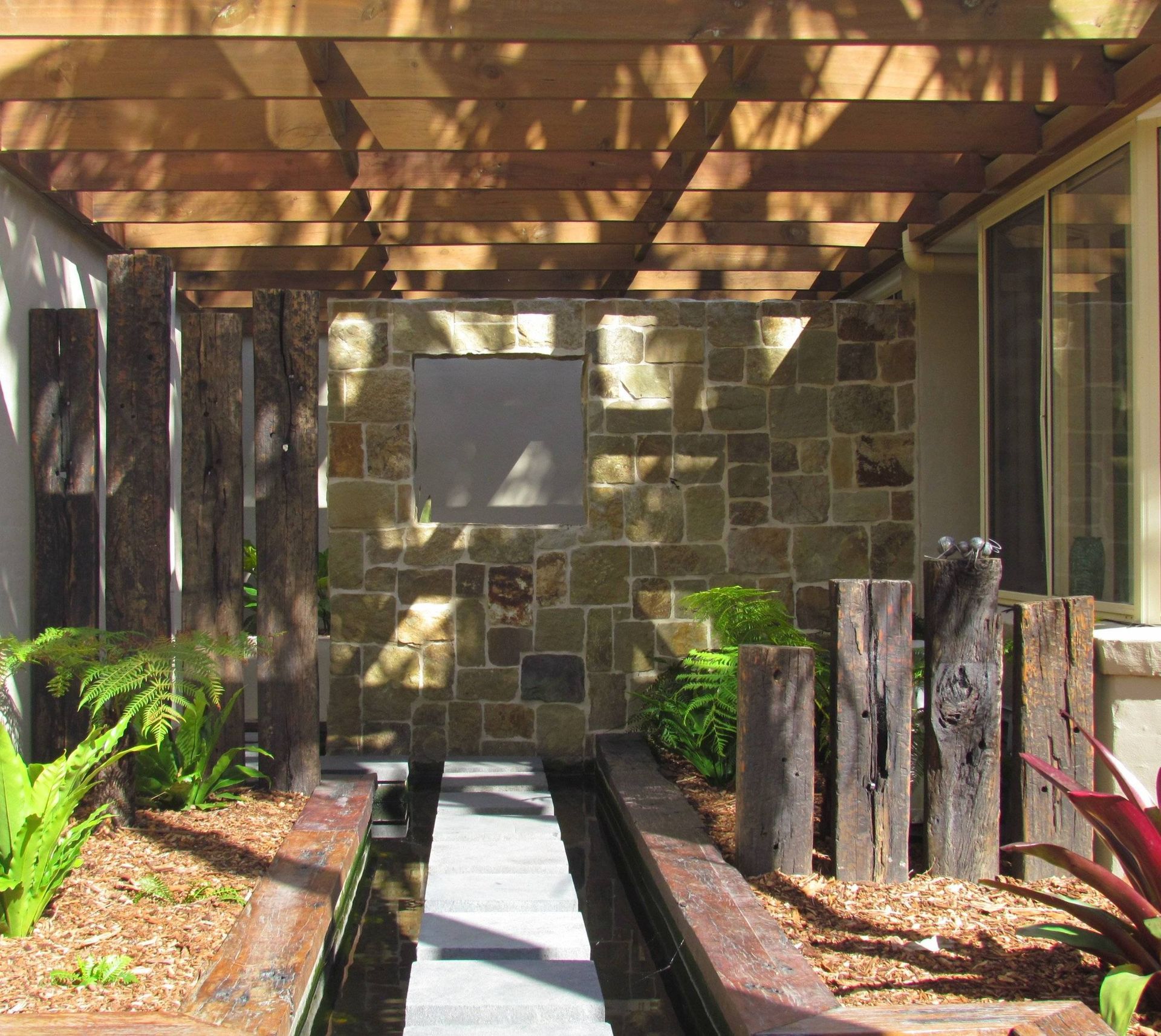 Wooden pergola over a stone path and rock wall, surrounded by landscaped planters and dark wood posts.