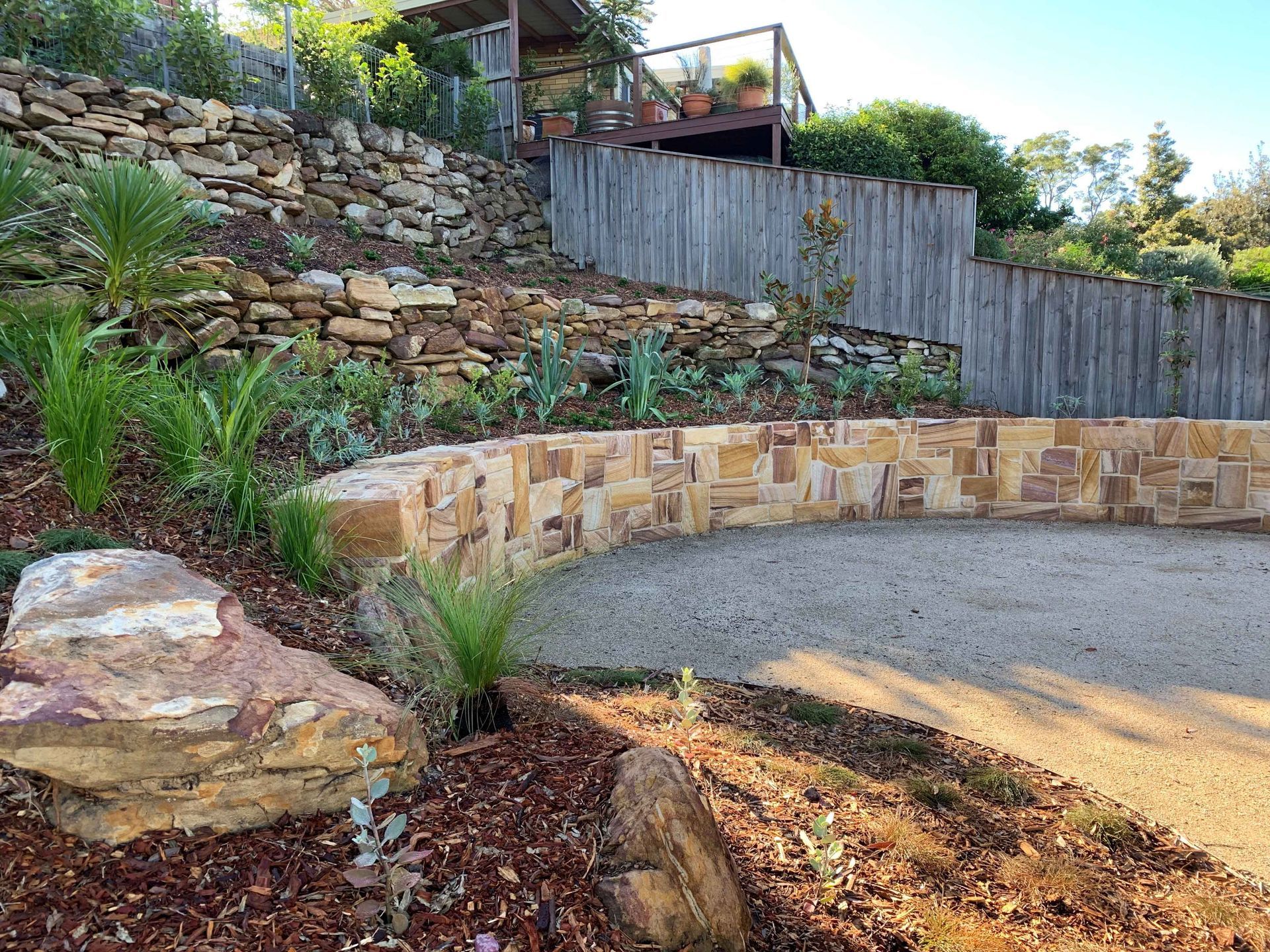 Stone retaining walls and a gravel driveway on a hillside, surrounded by native plants.