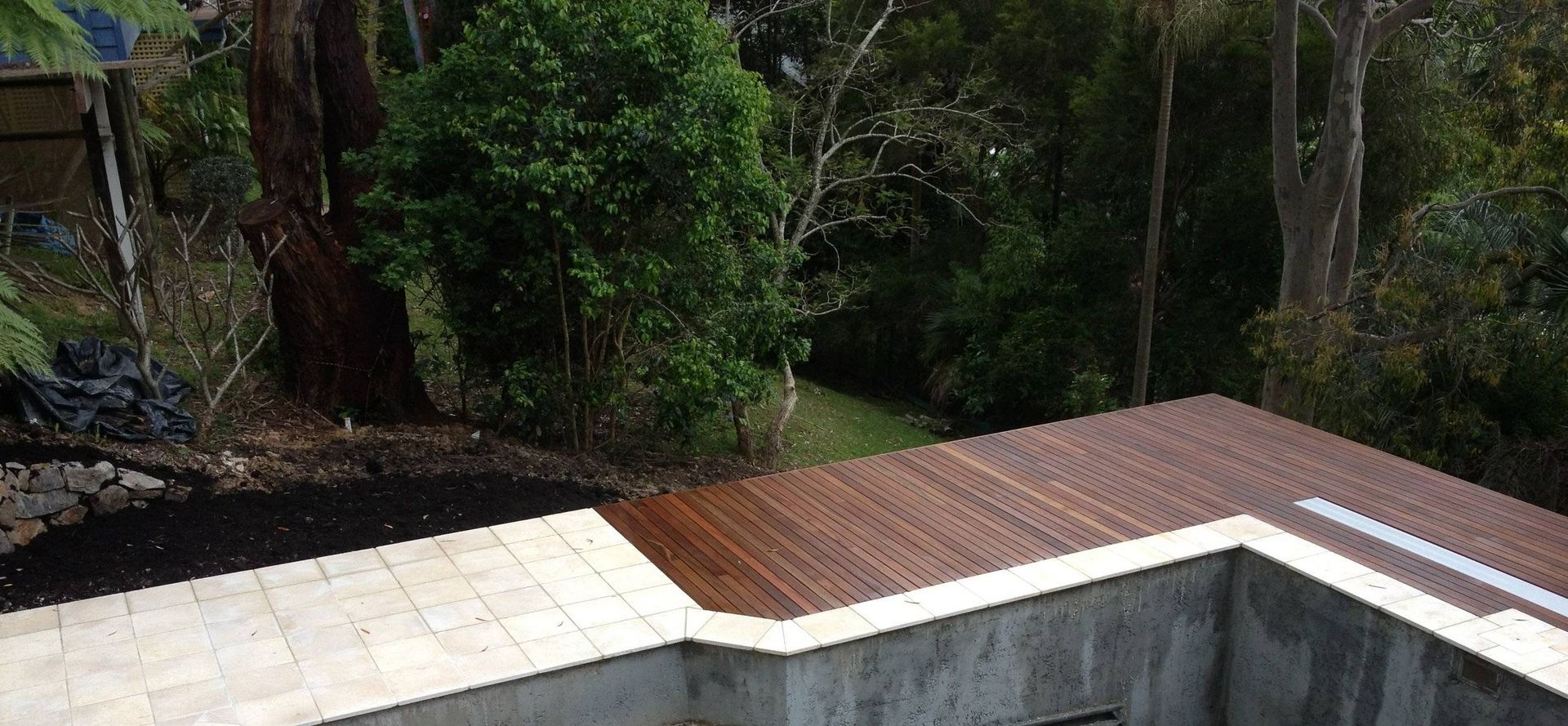 A wooden deck with a forest in the background. White stone tiles and a concrete wall are also visible.