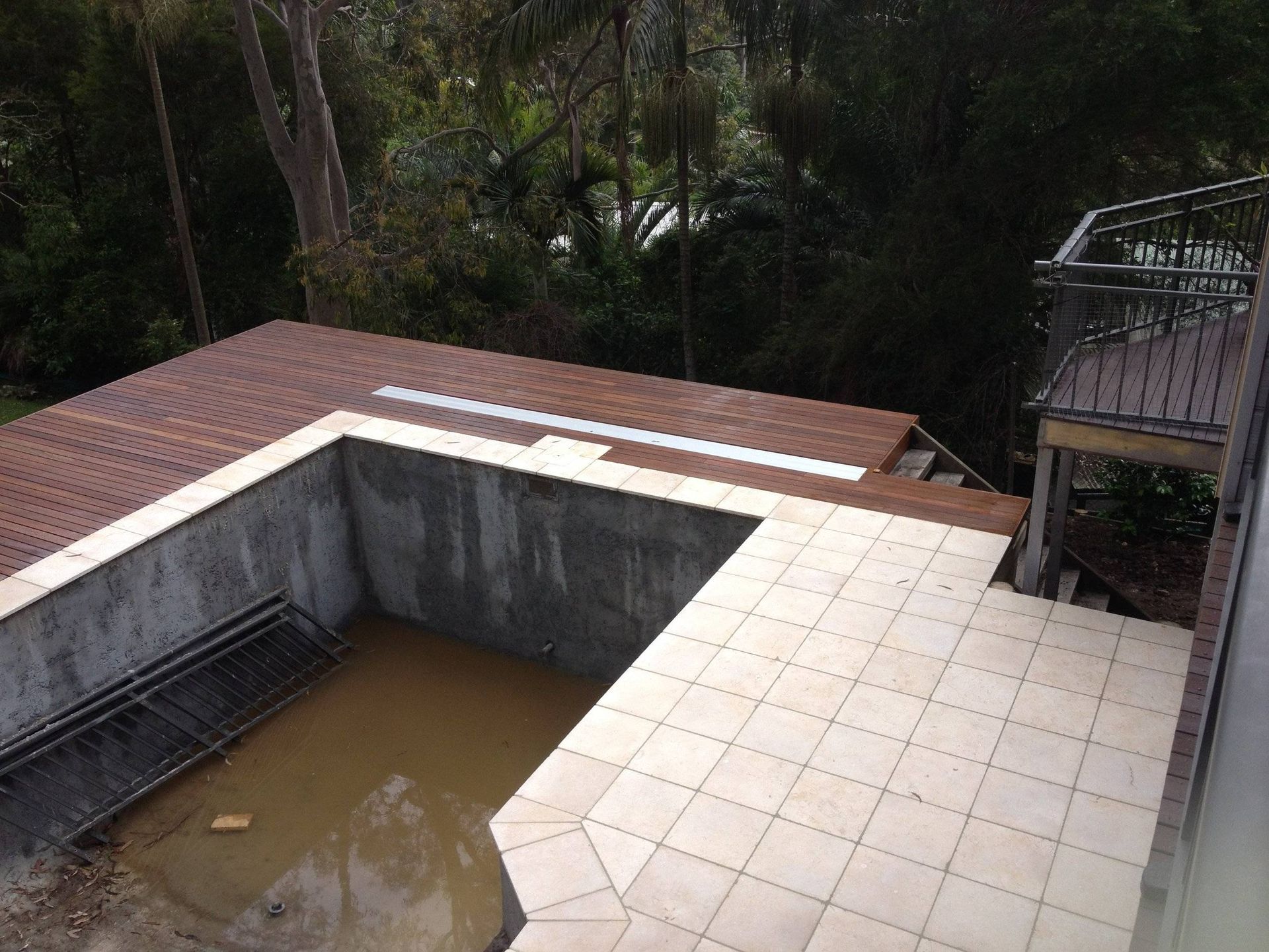 Partially built swimming pool, brown tiles, tan deck, concrete walls, surrounded by trees.
