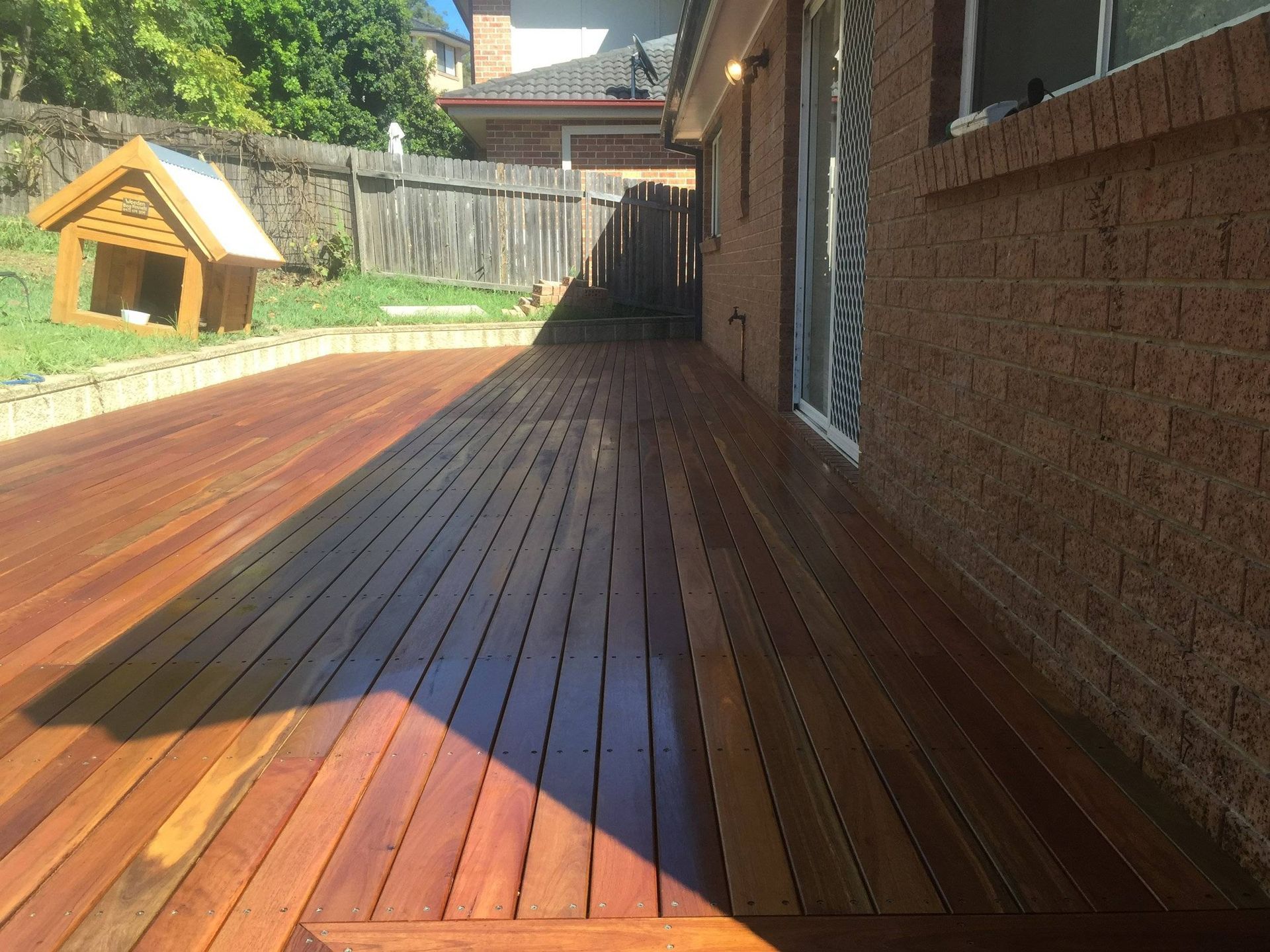 Wooden deck next to a brick building and a fenced backyard, with a dog house.