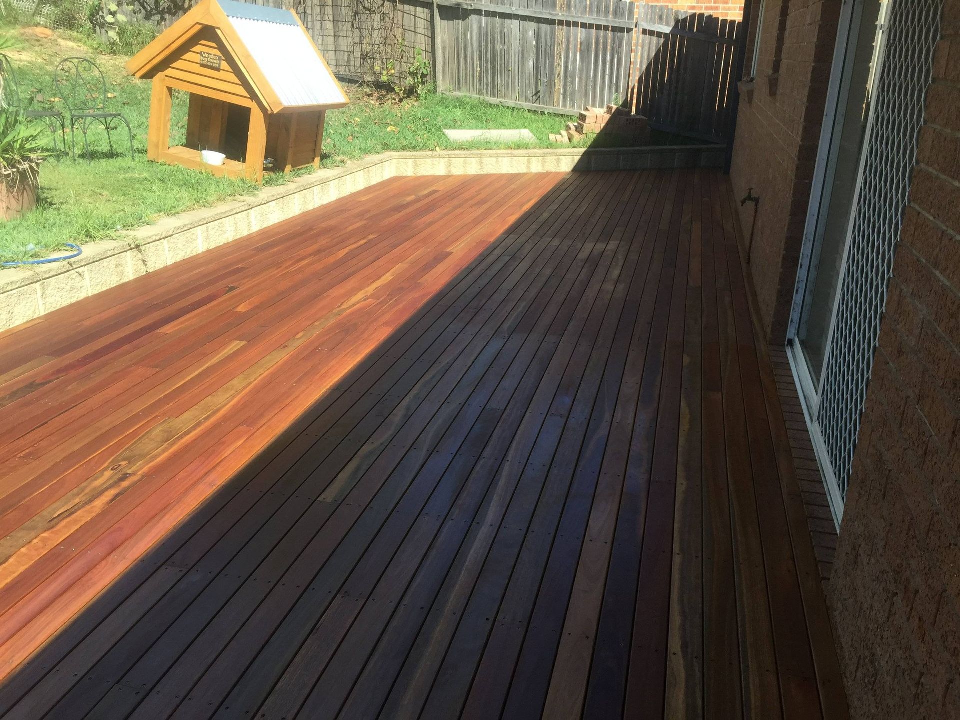 Wooden deck with a dog house on the grass, next to a brick building, casting long shadows.