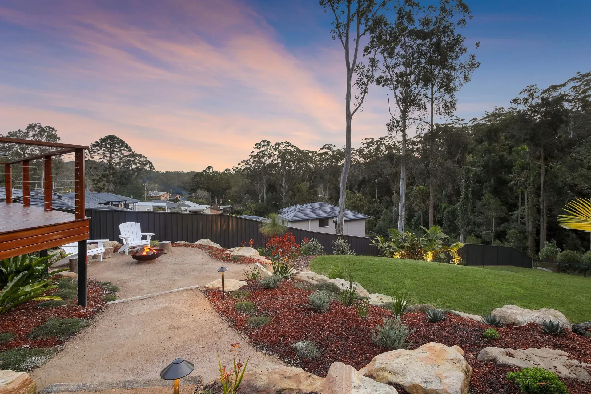 Backyard with fire pit, stone path, landscaping, and deck against a twilight sky with trees.