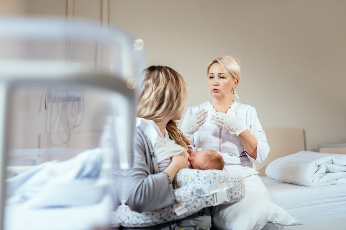 A woman is breastfeeding a baby in a hospital room while a nurse looks on.