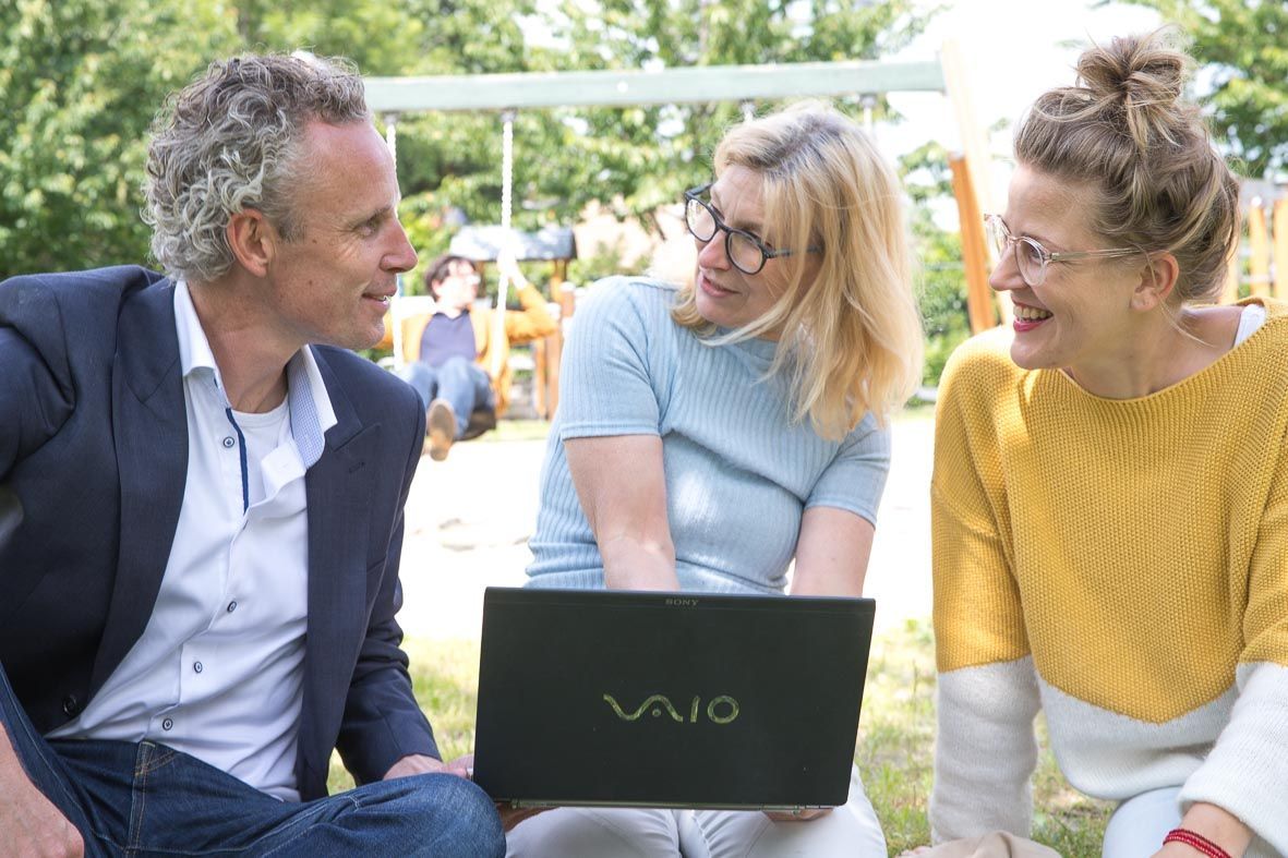 Drie mensen zitten in het gras en kijken naar een laptop. Op de achtergrond staat een schommel.
