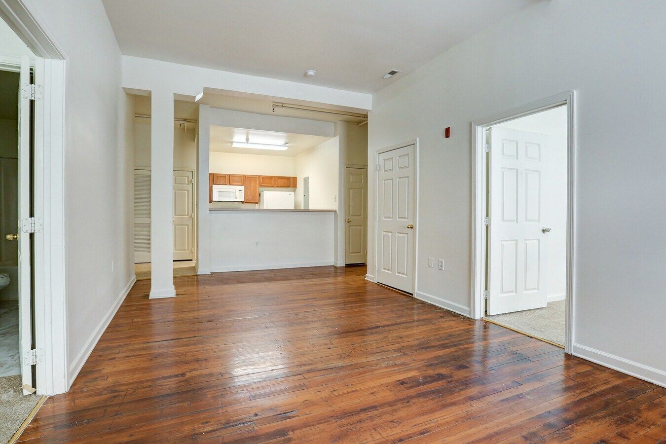 Empty apartment interior with wood floors, white walls, and a view into the kitchen.