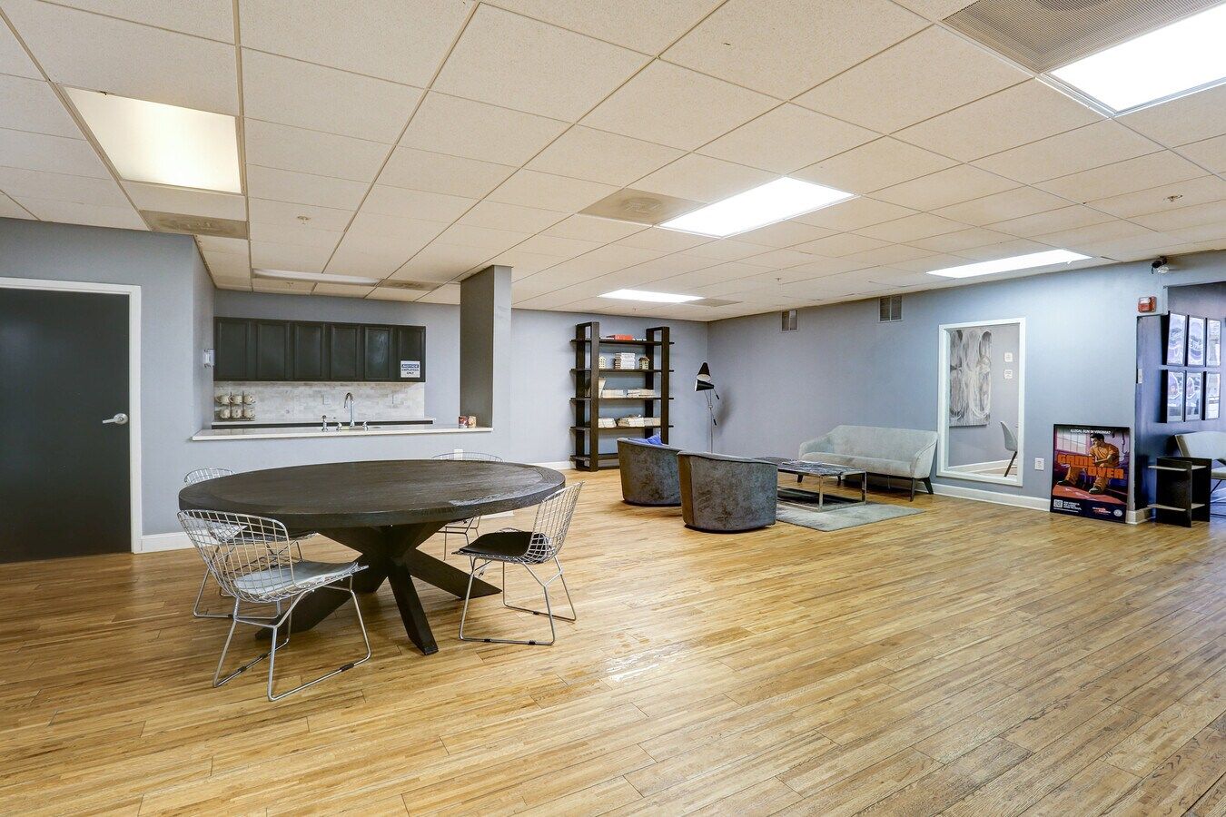 An open room with a round table and chairs, kitchen area, and seating area, with wood floors and grey walls.