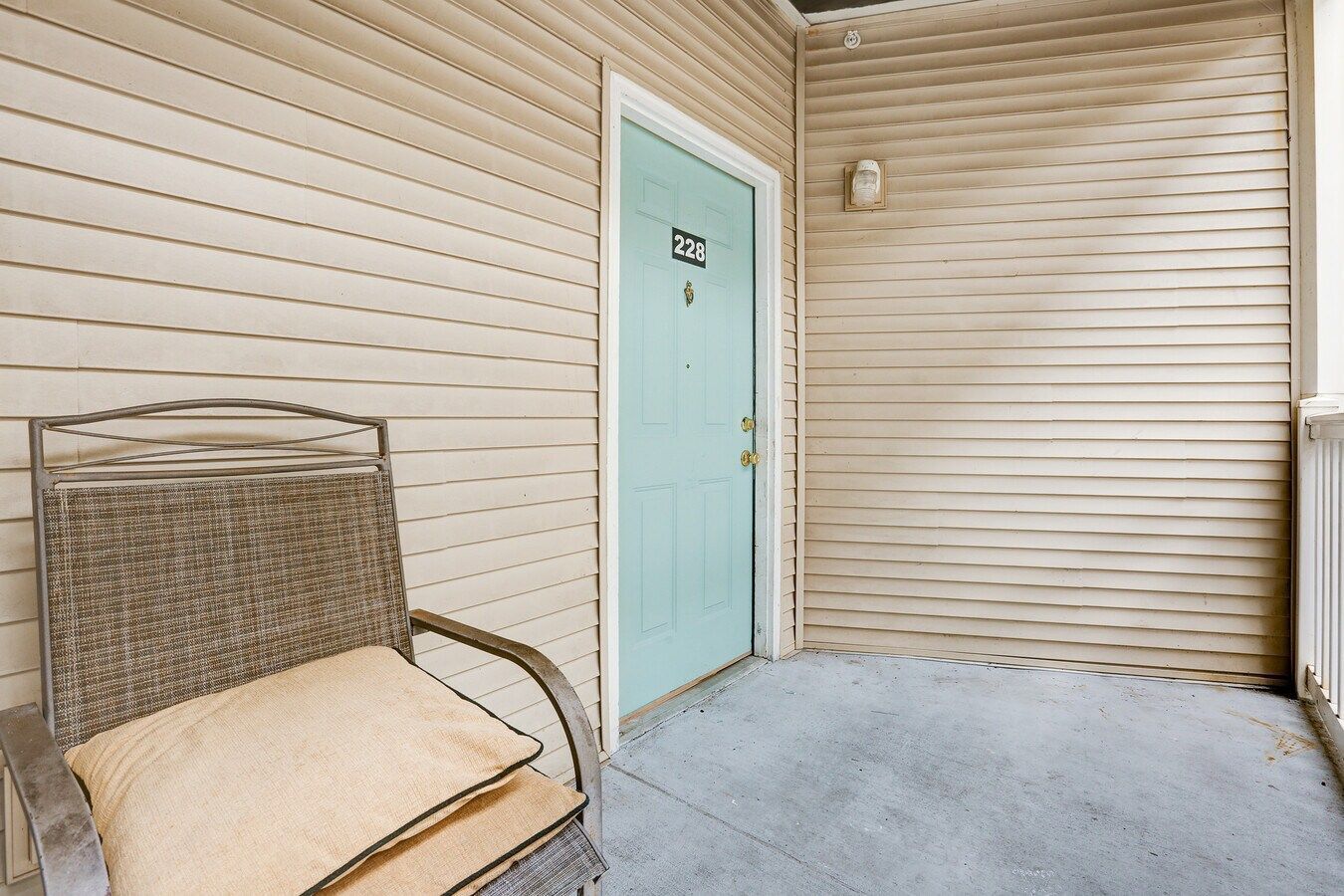A light blue door with the number 12 on it, on a beige-sided porch with a chair.