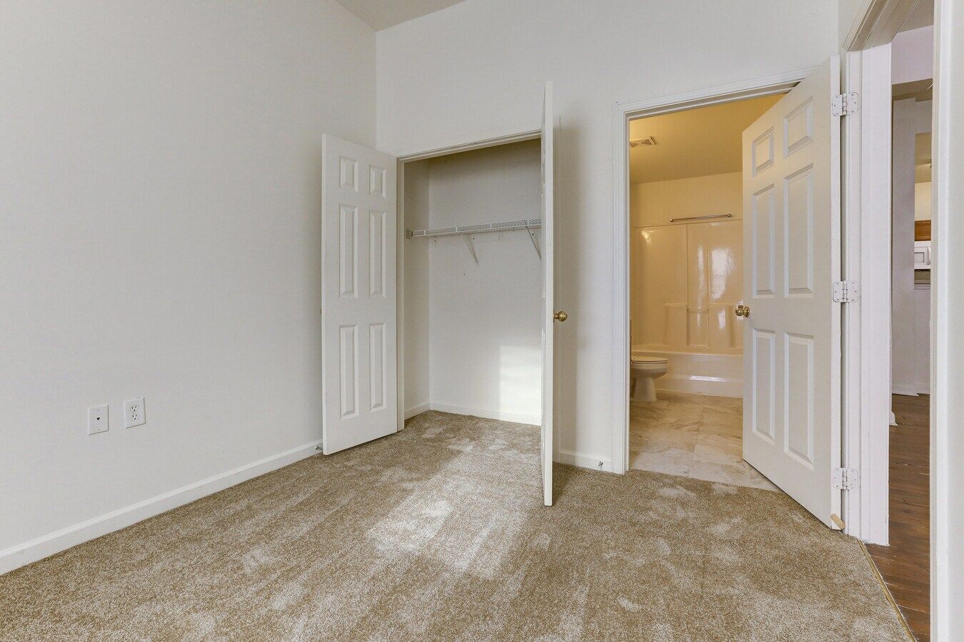 Empty bedroom with open closet and bathroom door. Beige carpet, white walls and doors.