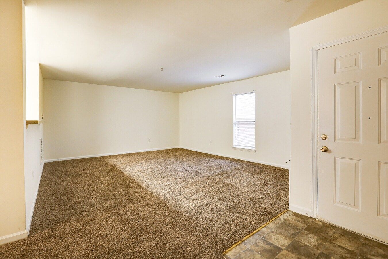 Empty living room with brown carpet, white walls, and a front door.