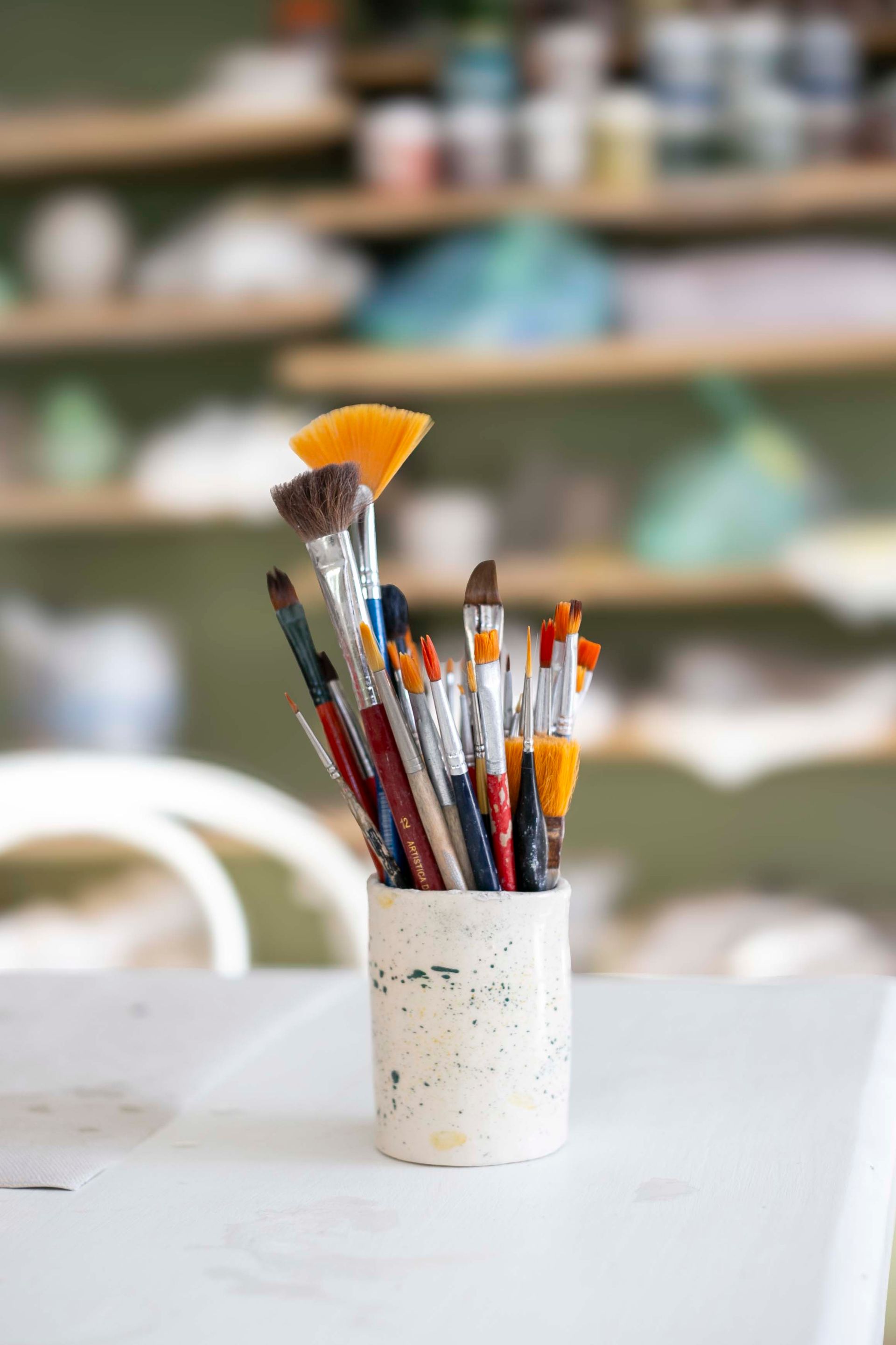 Pinceles en un recipiente de cerámica sobre una mesa blanca; estantes con materiales de arte en el fondo.