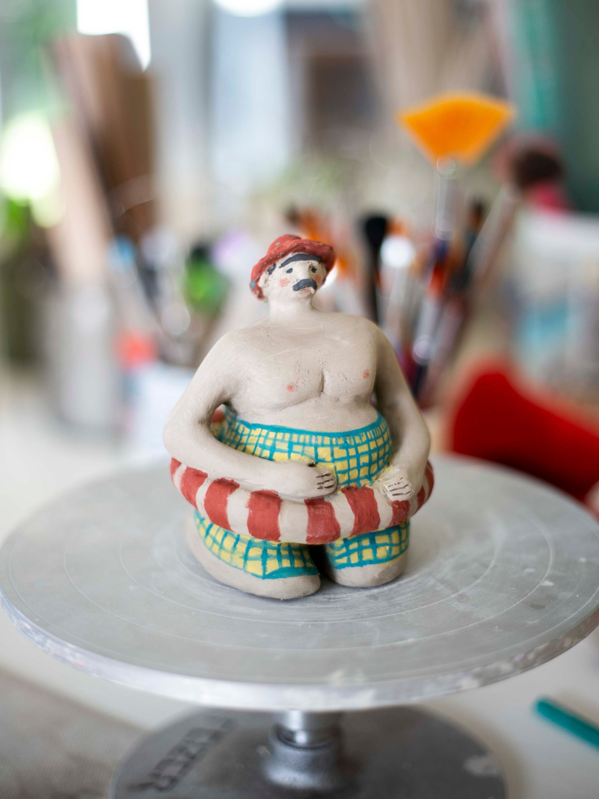 Figura de cerámica de una persona en traje de baño y sombrero, sosteniendo un salvavidas; sobre un torno de alfarero.