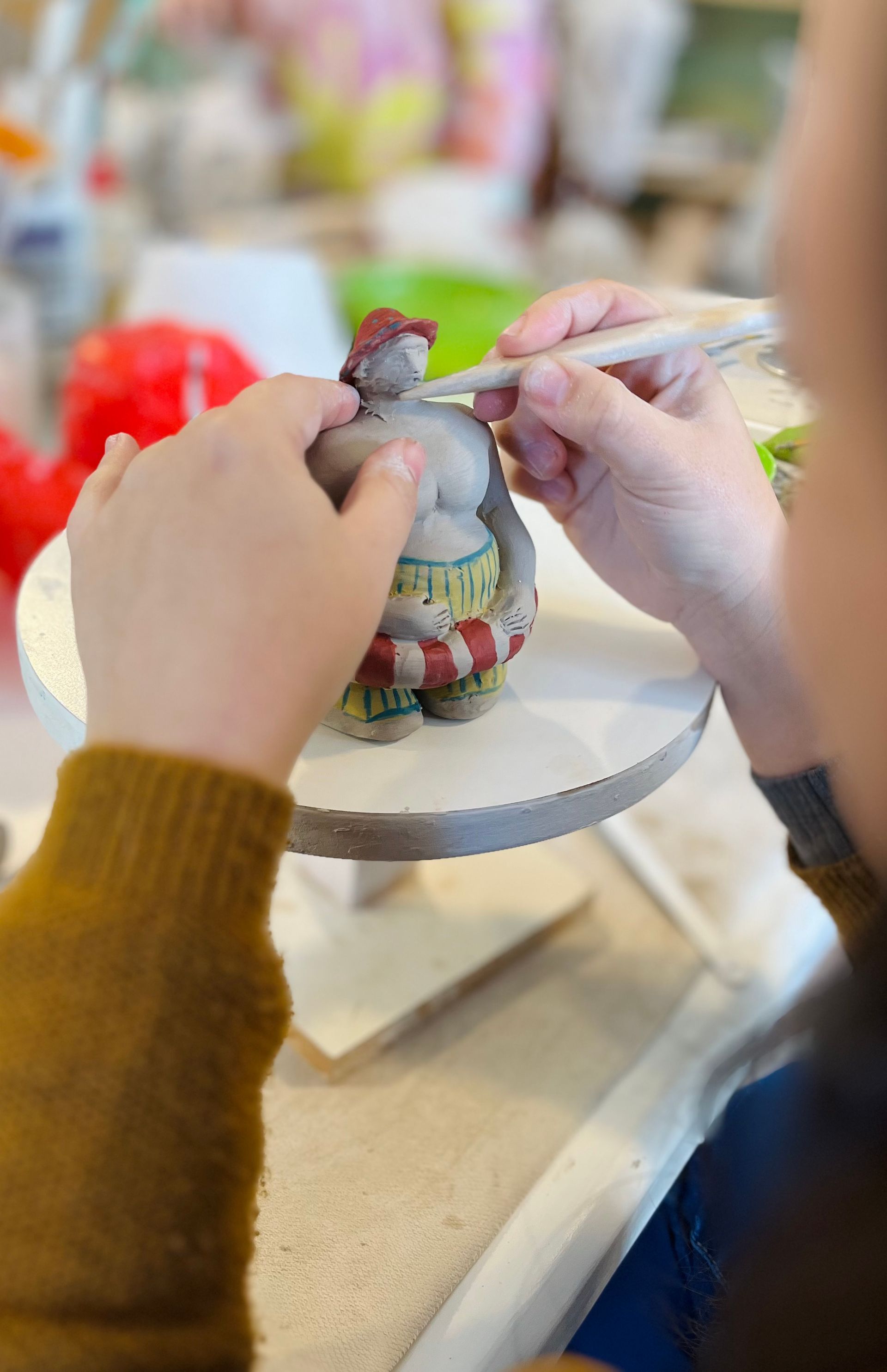 Persona esculpiendo una figura de arcilla con una herramienta; la figura está en un torno de alfarero.