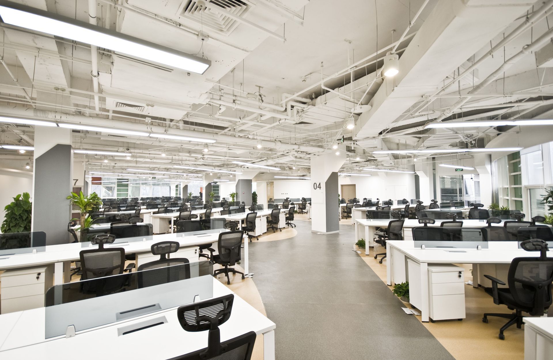 Open-plan office with rows of white desks, black chairs, computers, and bright ceiling lights