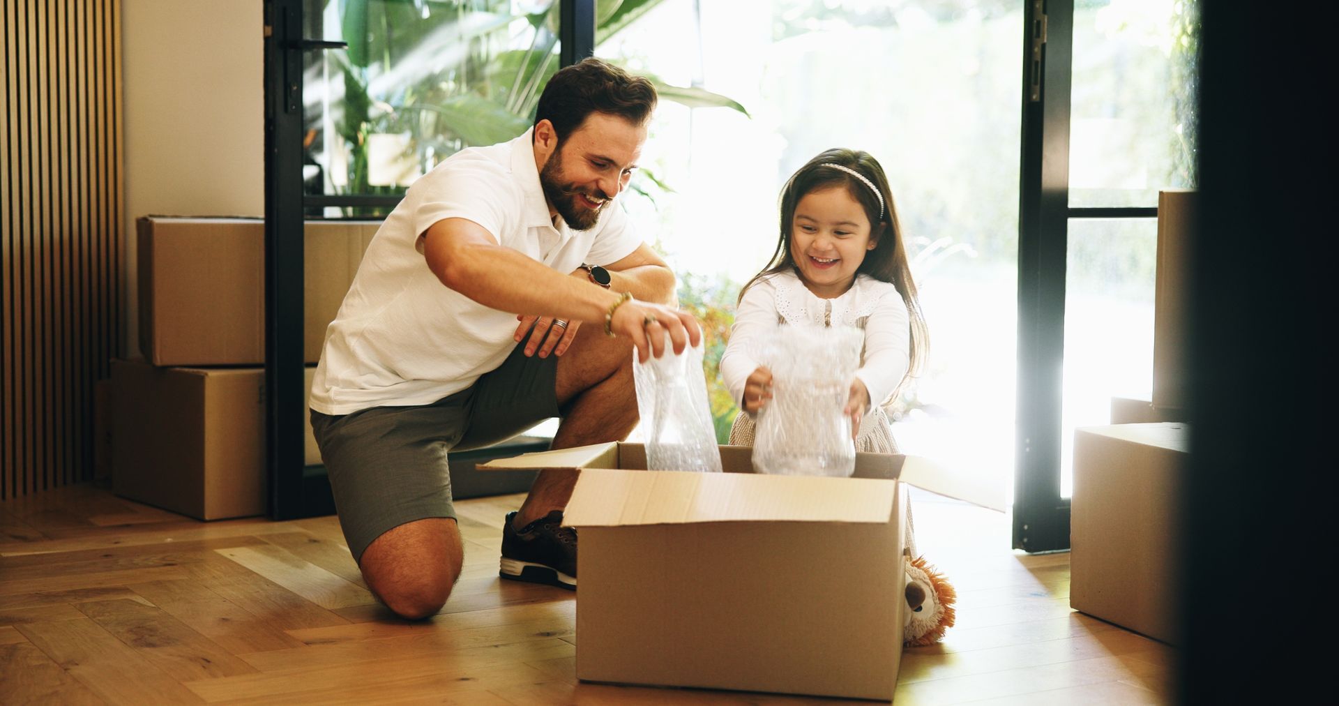 Man and child unpacking a box in a sunlit room, smiling near a large open window.