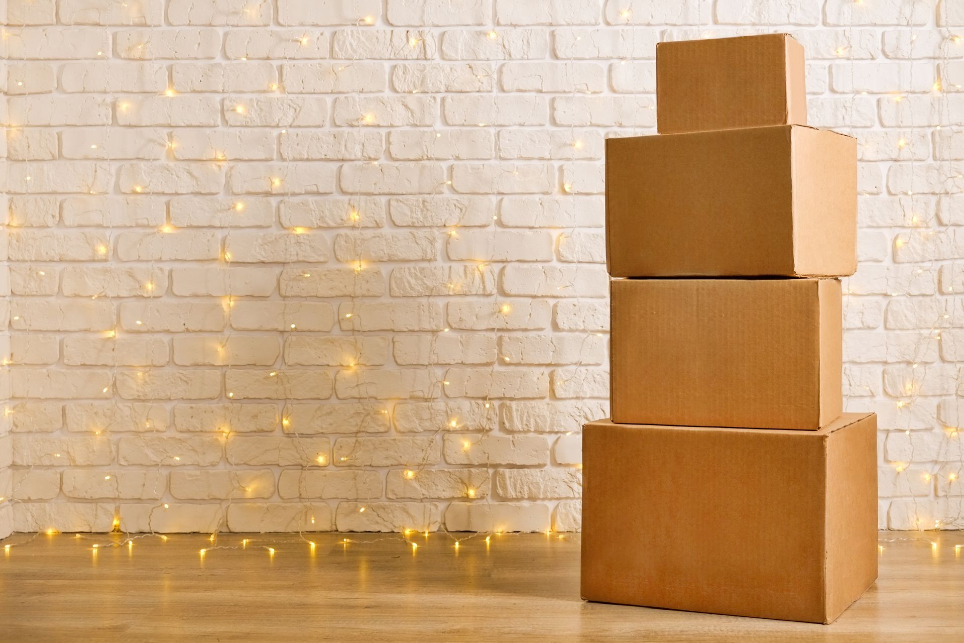 Stack of four cardboard boxes against a white brick wall with string lights on a wooden floor.