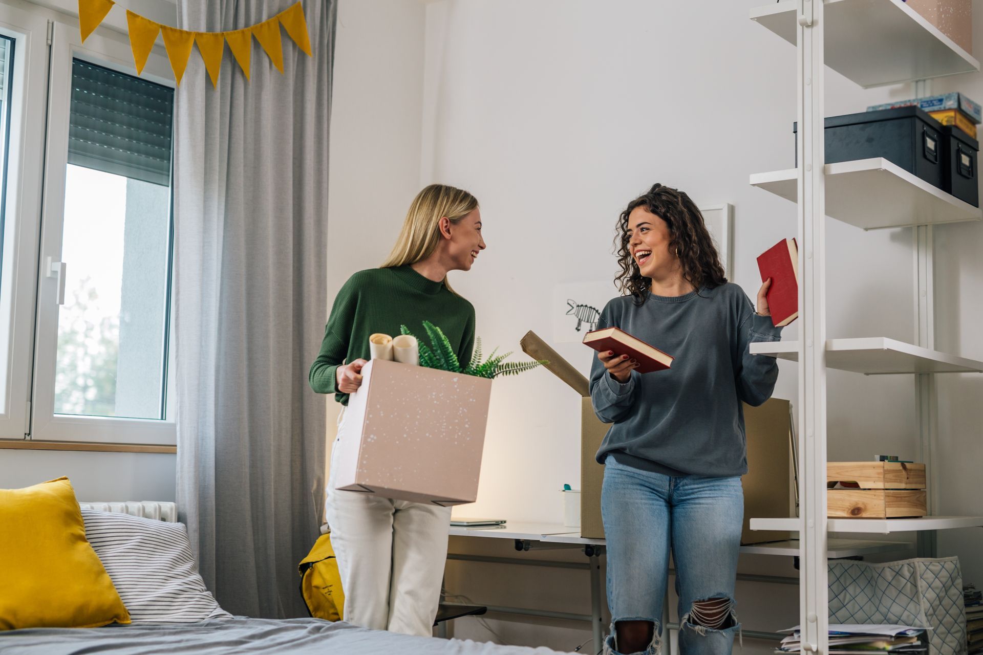 Two women unpacking boxes in a bright room with a bed and shelves. One holds a book, the other a box.