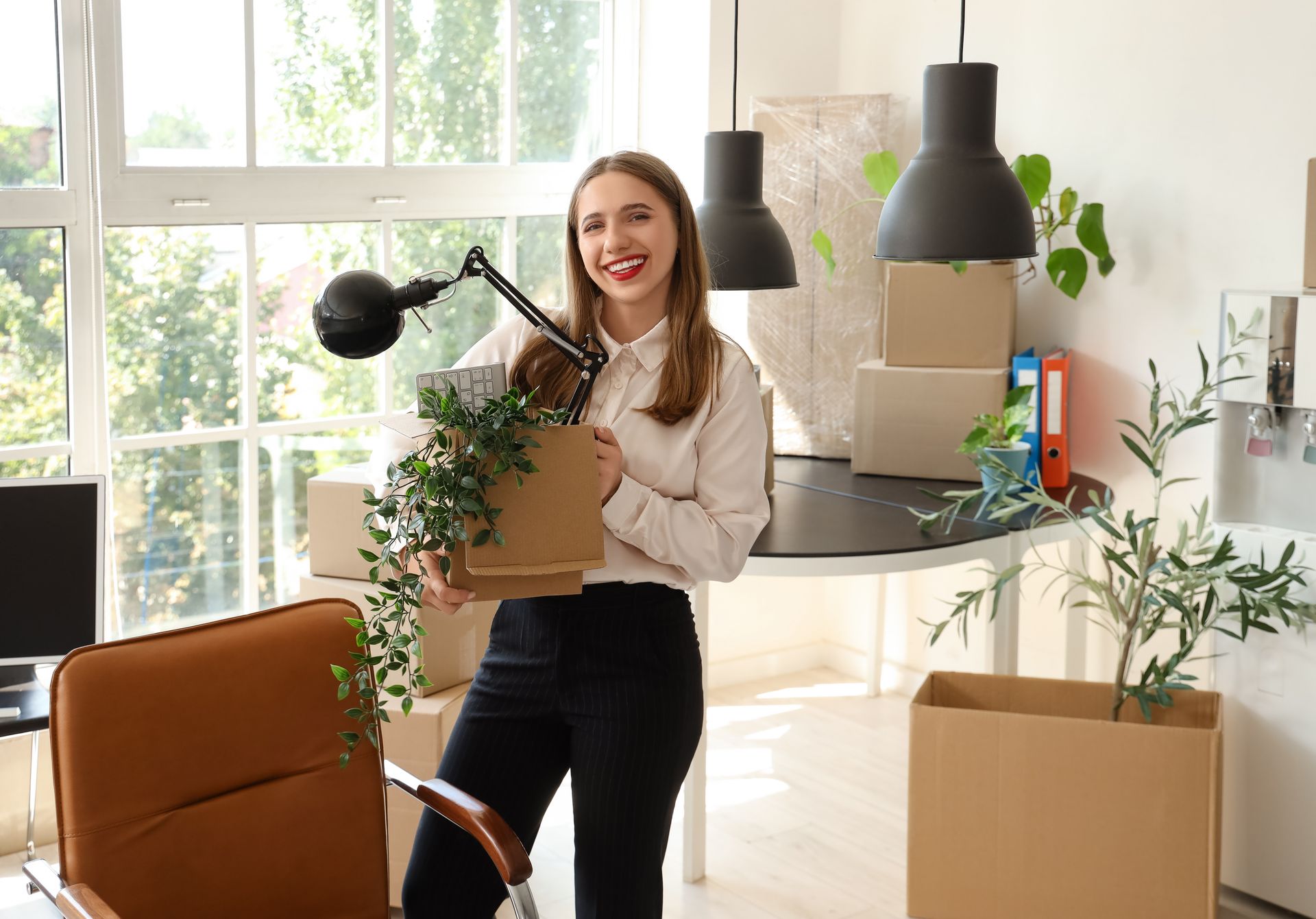 Smiling woman carrying a box and lamp in a bright living room with moving boxes and plants