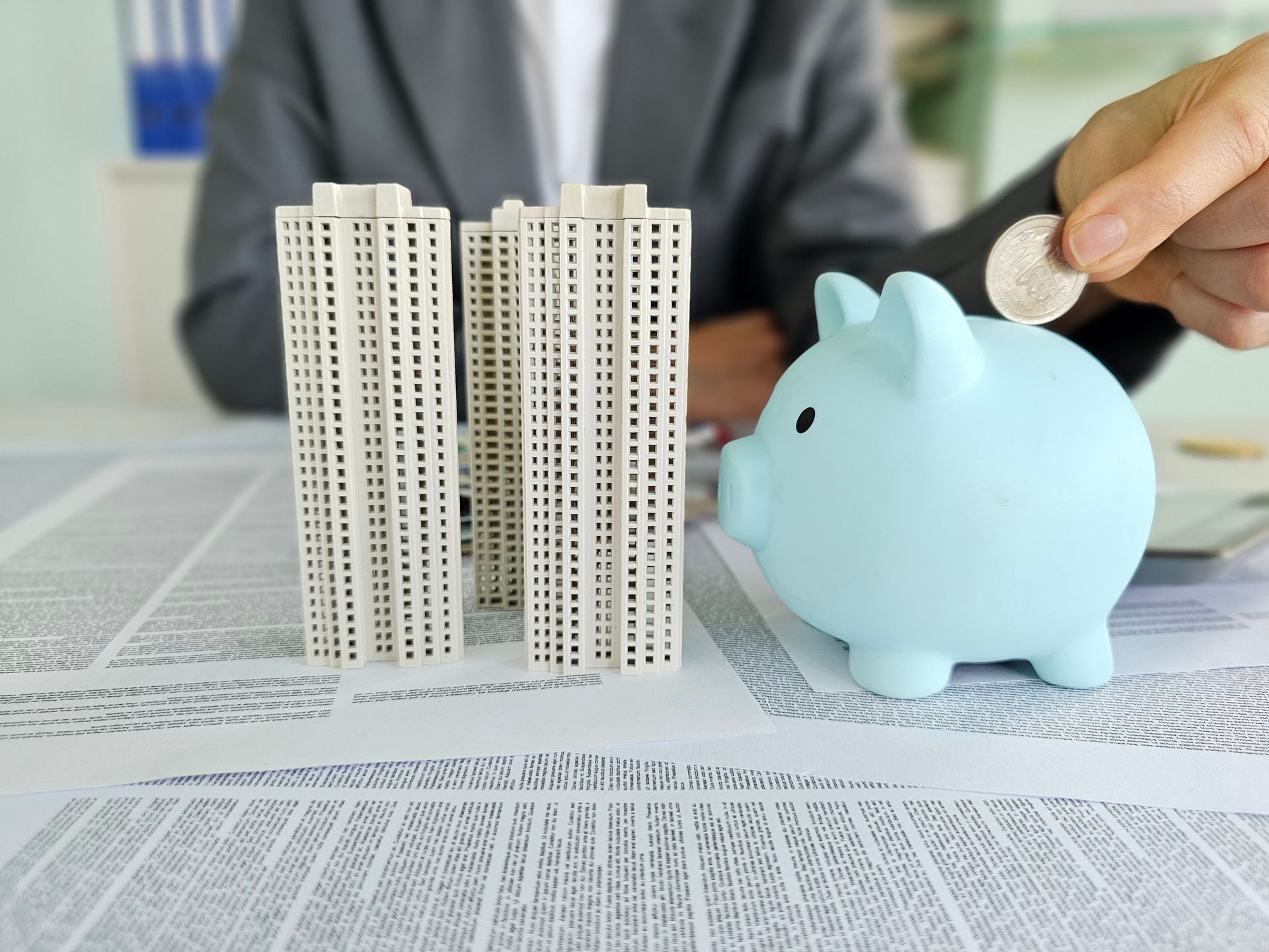 Person putting coin into a blue piggy bank near model buildings on a desk.