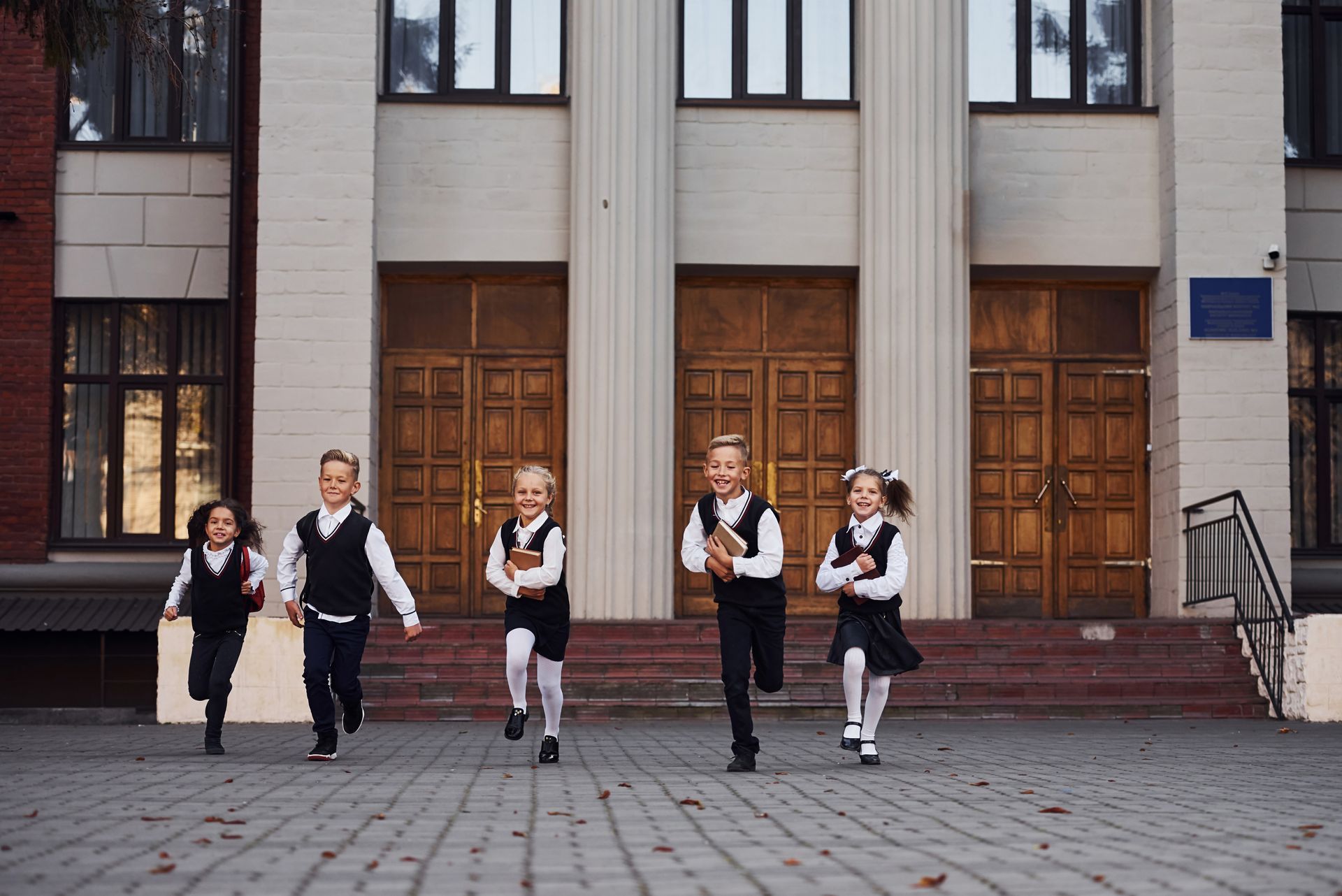 A group of children in school uniforms are running in front of a school building.