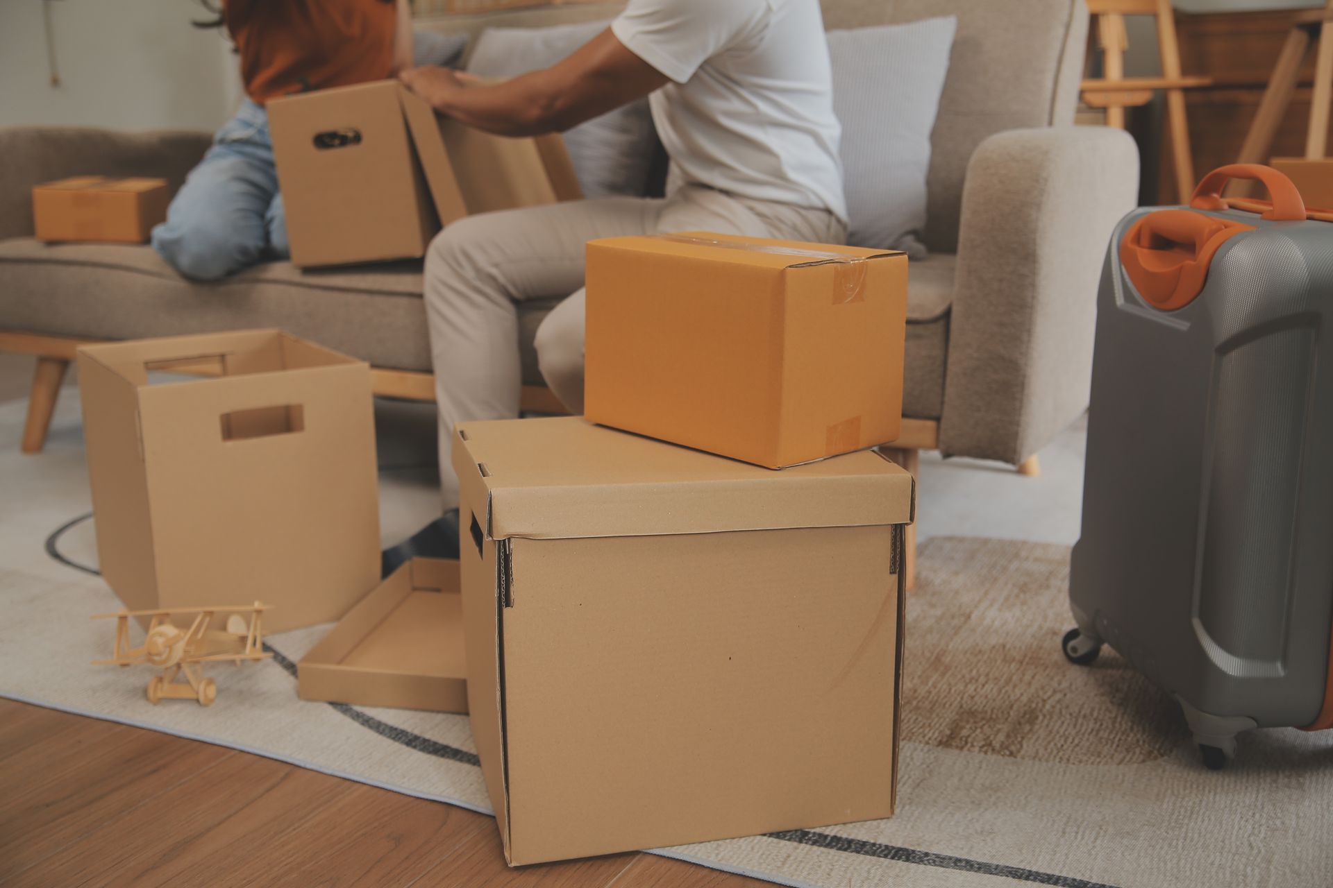 A man is sitting on a couch surrounded by cardboard boxes and suitcases.