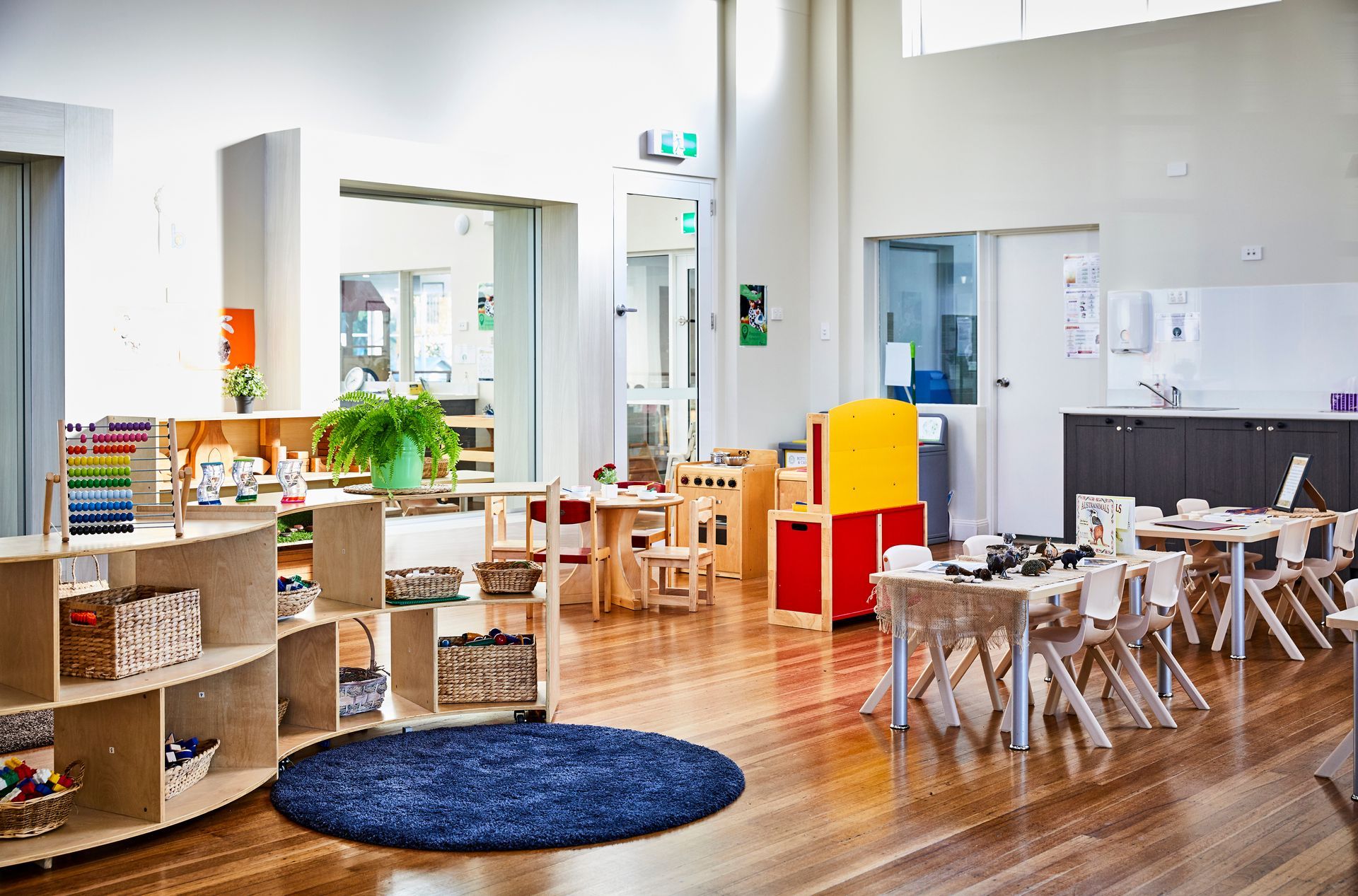 Bright preschool classroom with wooden tables, toys, plants, and a blue rug on a polished floor