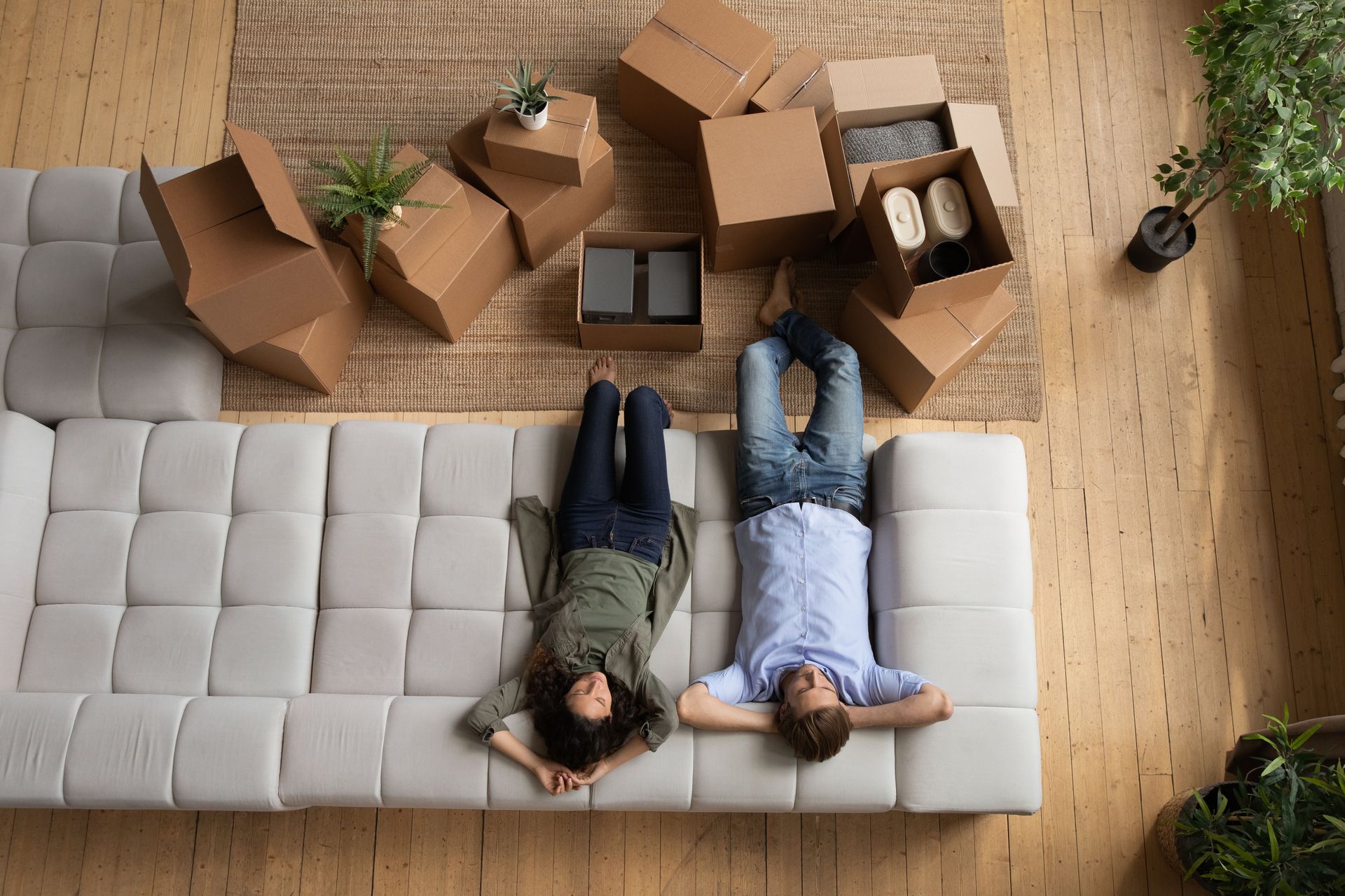 A man and a woman are laying on a couch in a living room surrounded by cardboard boxes.