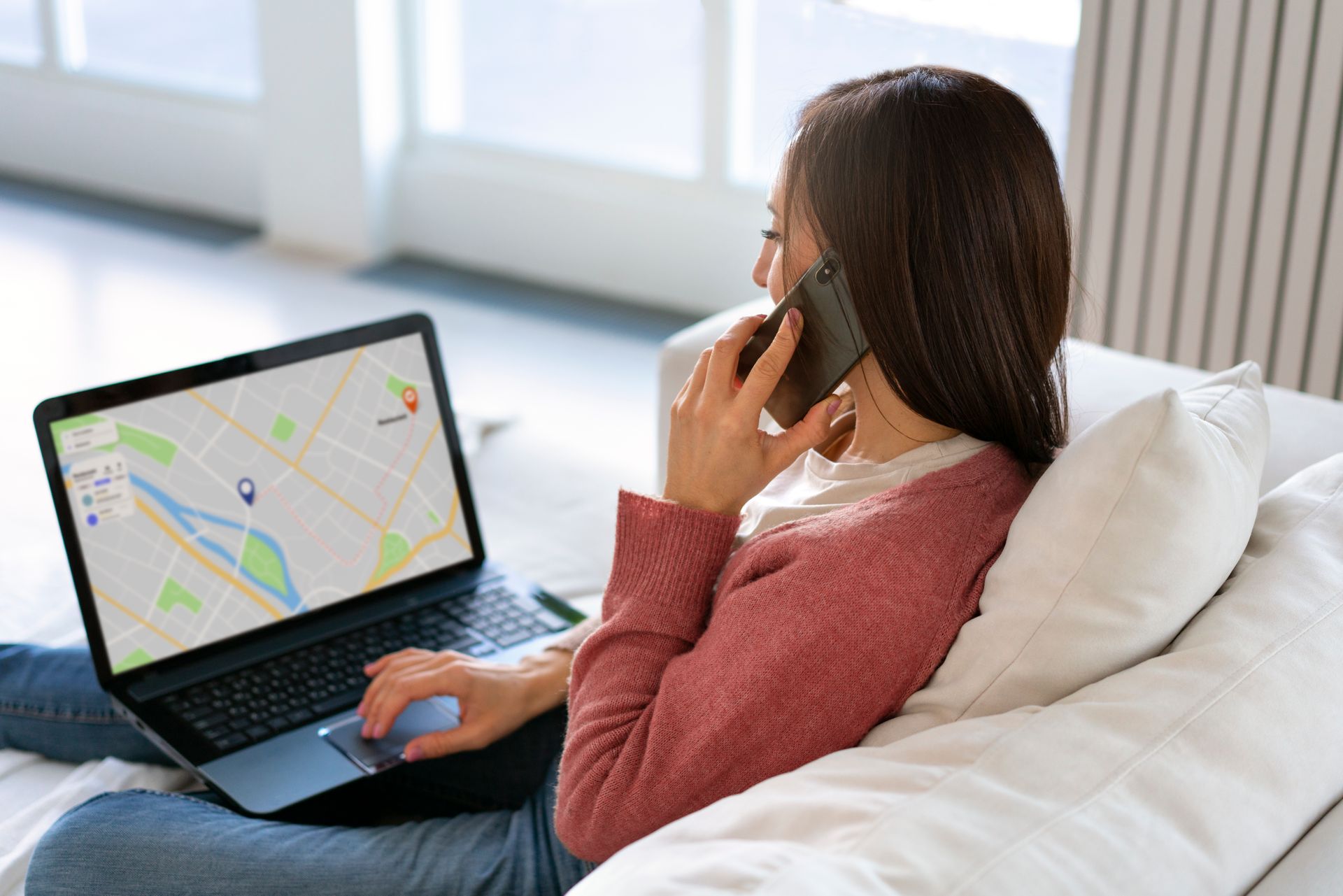 A woman is sitting on a couch using a laptop and talking on a cell phone.