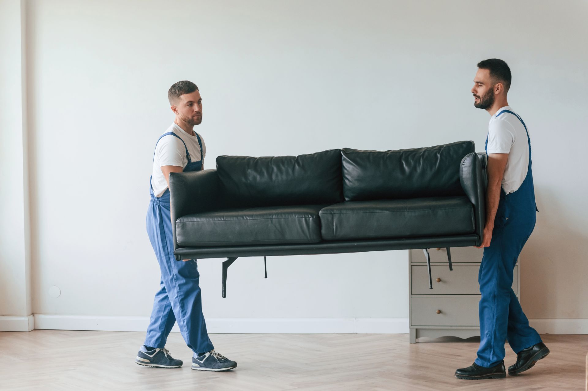 Two men are carrying a black couch in a living room.