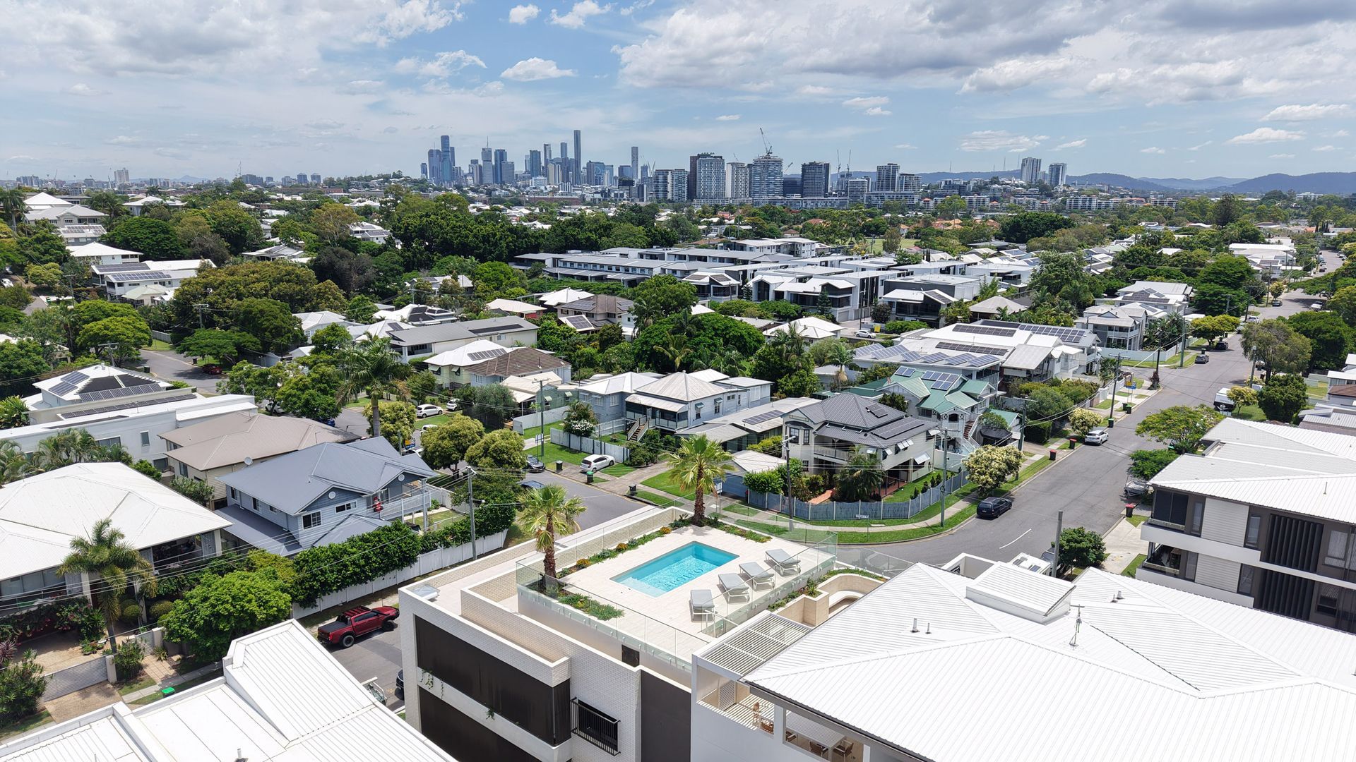 Aerial view of a residential area with a city skyline in the distance. A building with a rooftop pool is in the foreground.