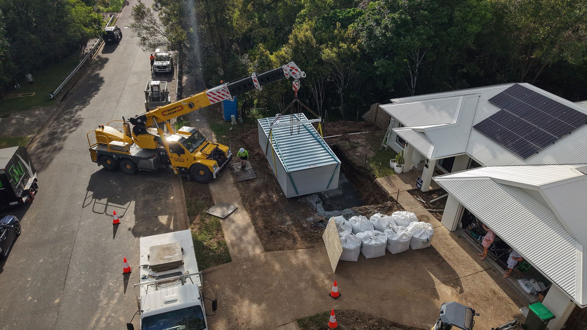 A crane lowers a large container onto a prepared site near a house with solar panels. Construction workers 