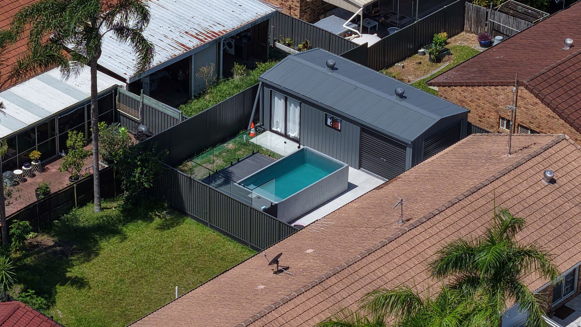 Aerial view of a small, modern house with a rectangular pool in the backyard, surrounded by fences and other houses.