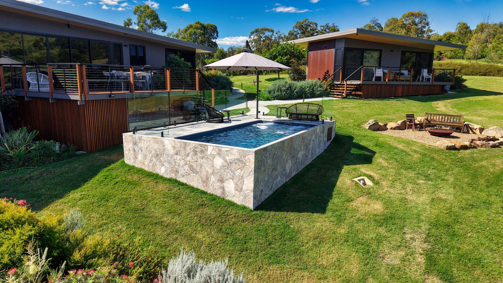 Stone-walled pool surrounded by green grass, with two cabins and an umbrella in the background.