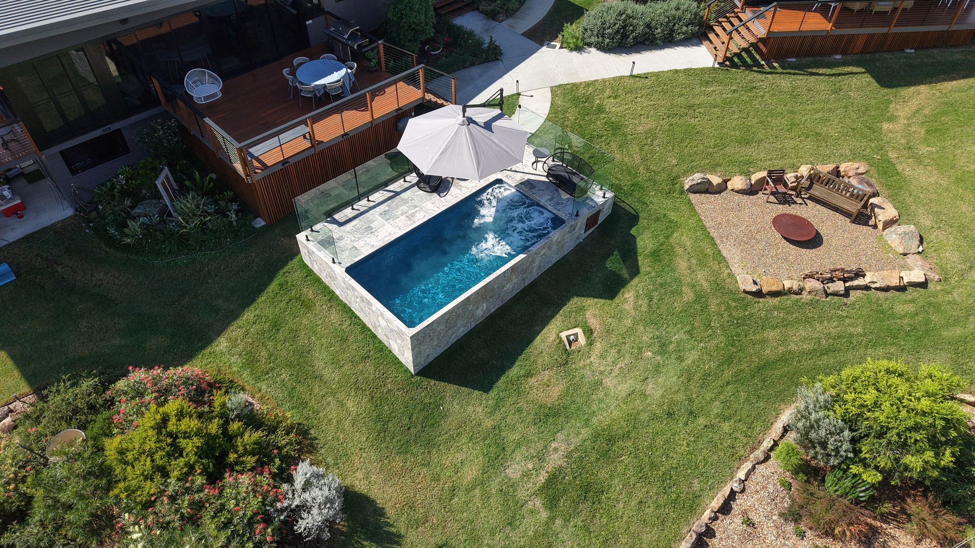 Aerial view of a backyard with a small pool, fire pit, and wooden deck, surrounded by green grass and landscaping.