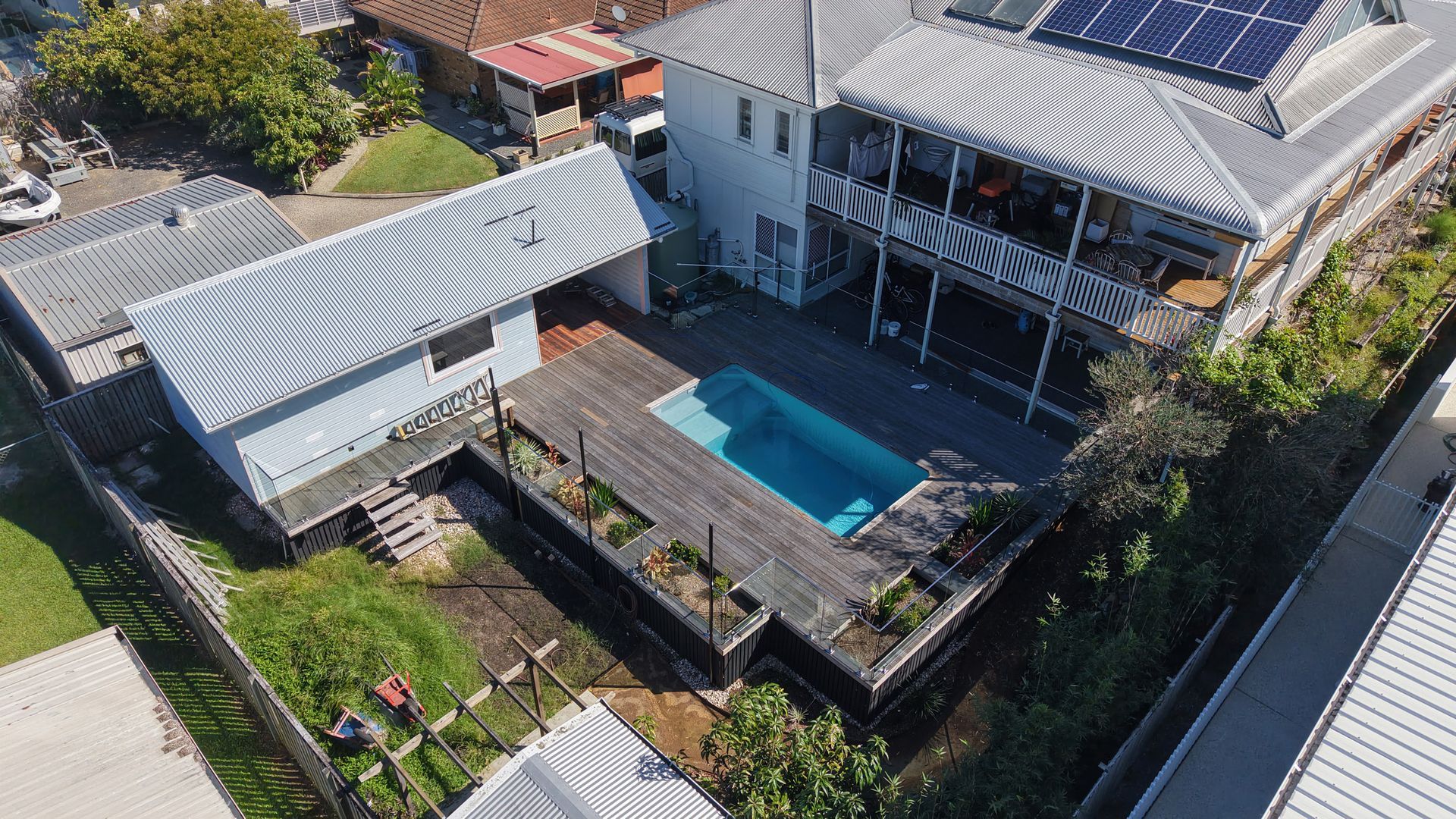 Aerial view of a home with a pool, wooden deck, and solar panels on the roof. The yard is surrounded by fencing and greenery.