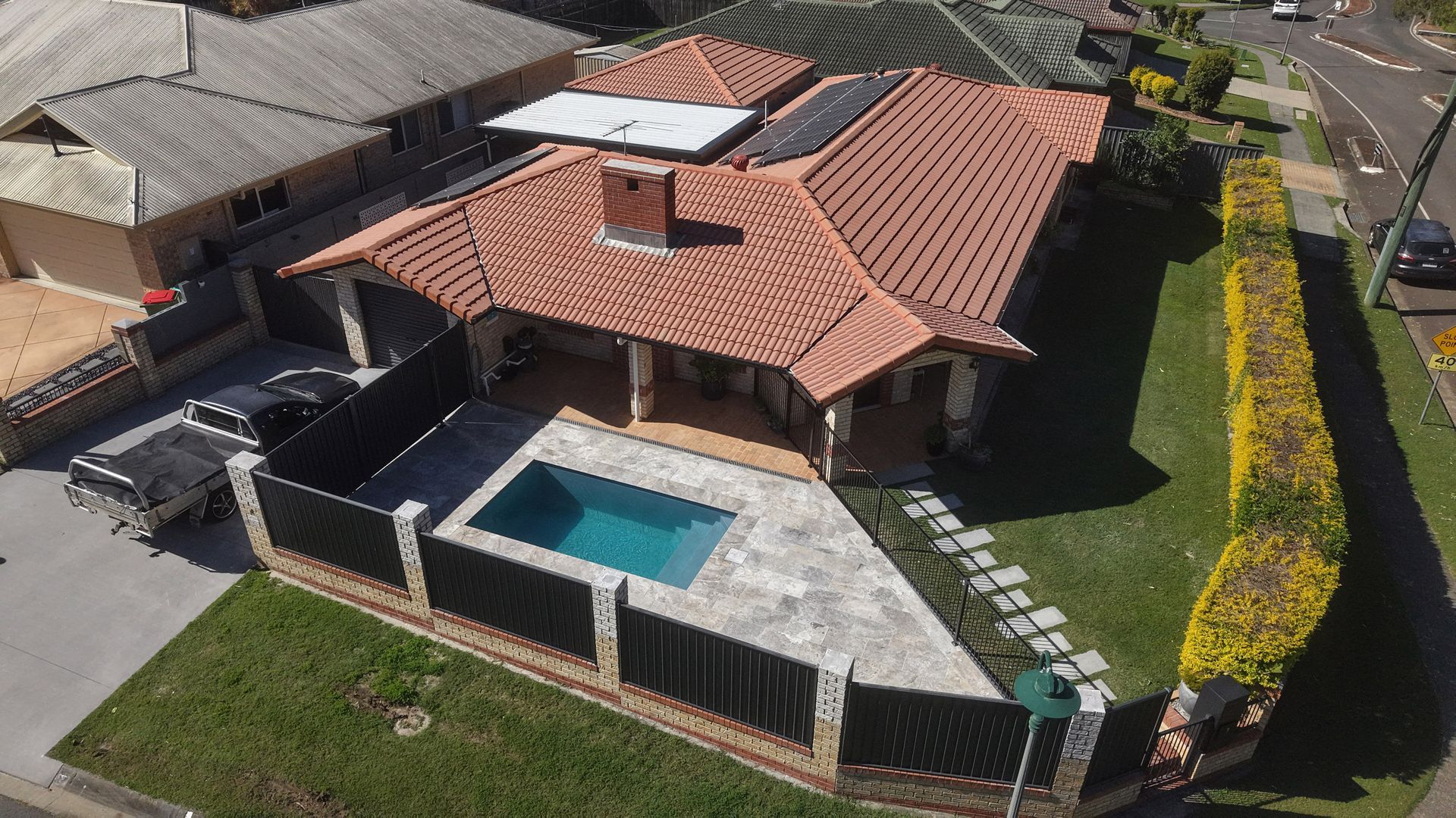 Aerial view of a house with a terracotta roof, a swimming pool, and a lush green lawn. A black fence surrounds the property.