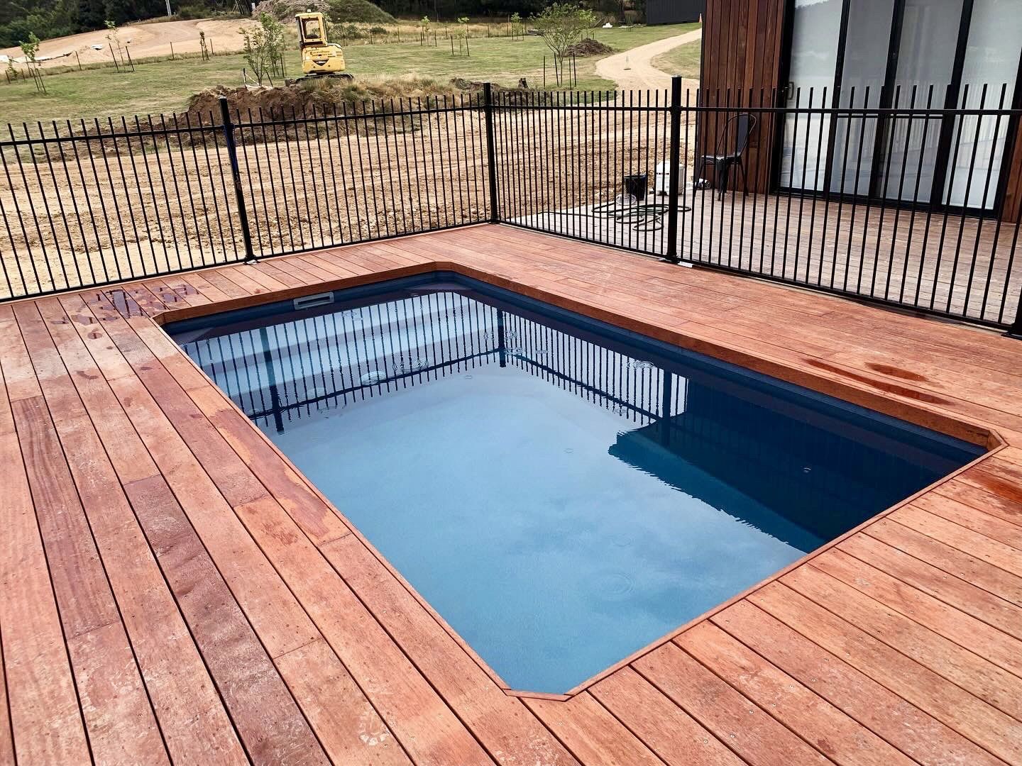 Rectangular plunge pool surrounded by reddish-brown wooden decking, enclosed by a black metal fence; a modern house