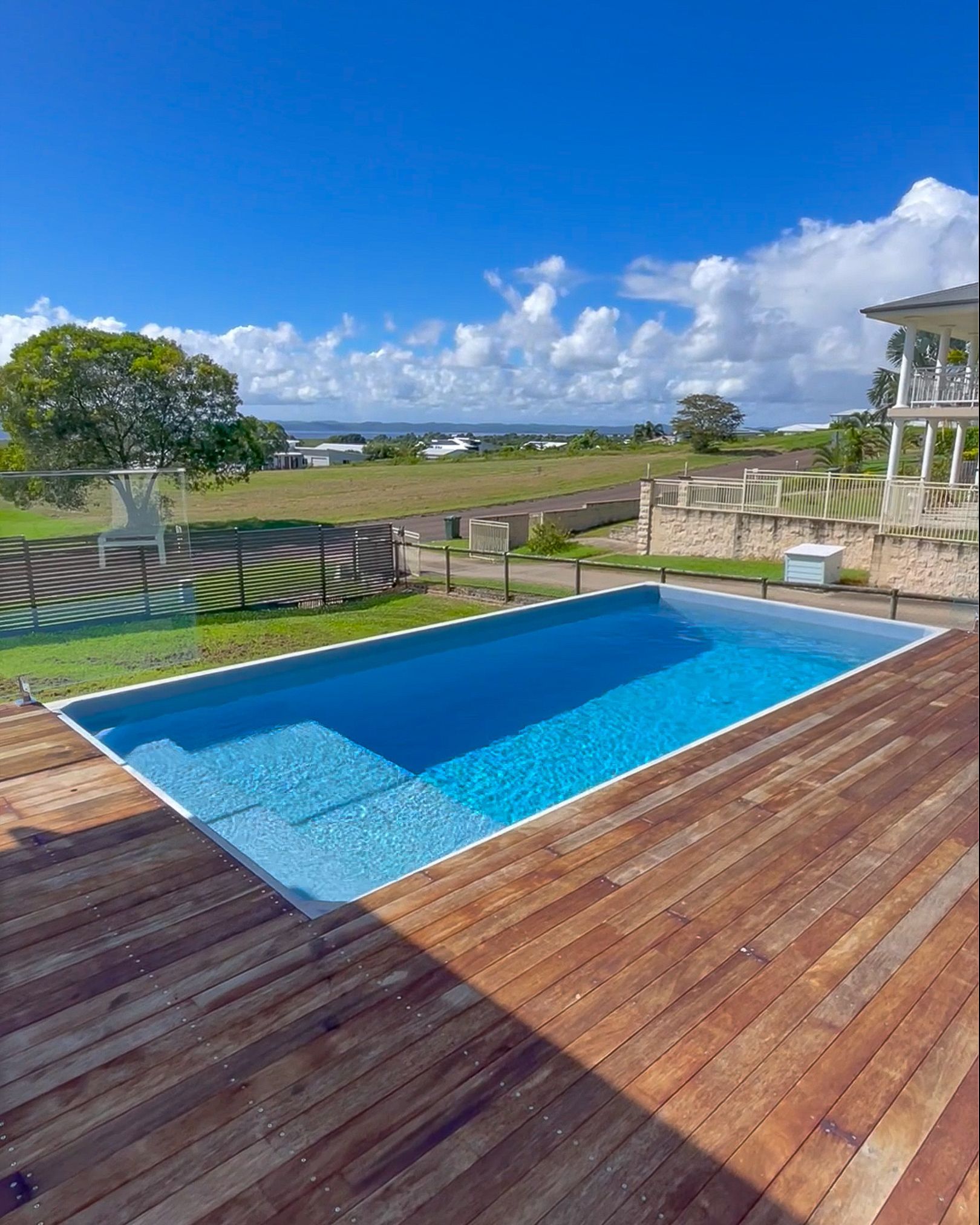Rectangular swimming pool with blue water surrounded by wooden decking, with a field, blue sky, and white clouds 