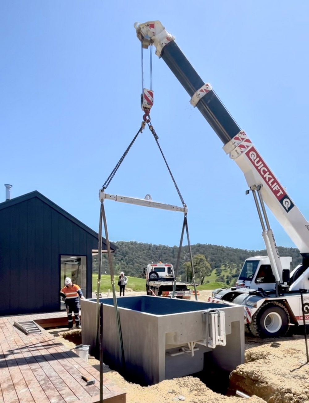 A crane lifts a gray concrete pool into place near a dark building. Workers in hard hats observe the installation.
