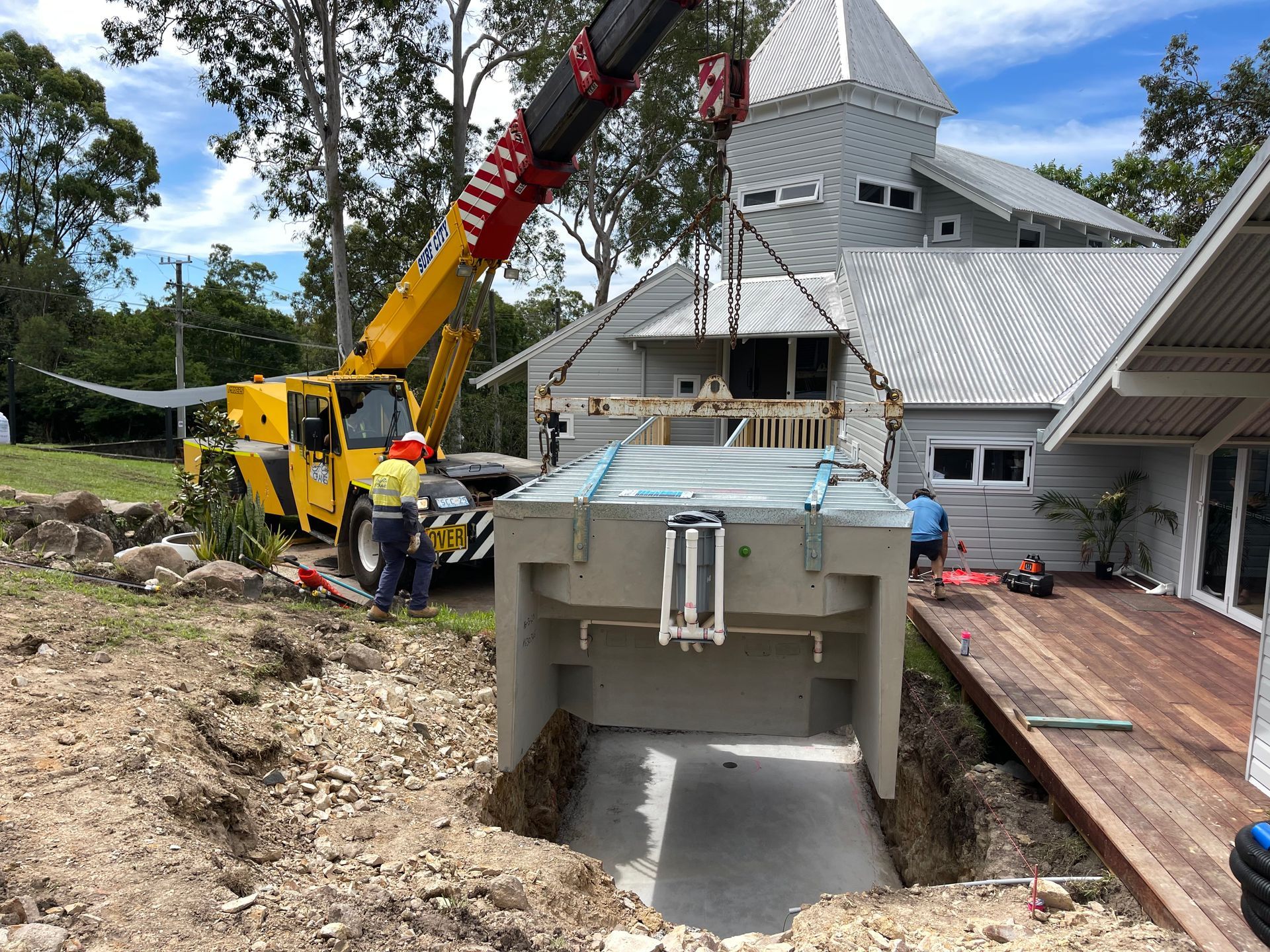 A crane lowers a large concrete tank into a trench near a house under construction. Two workers supervise.
