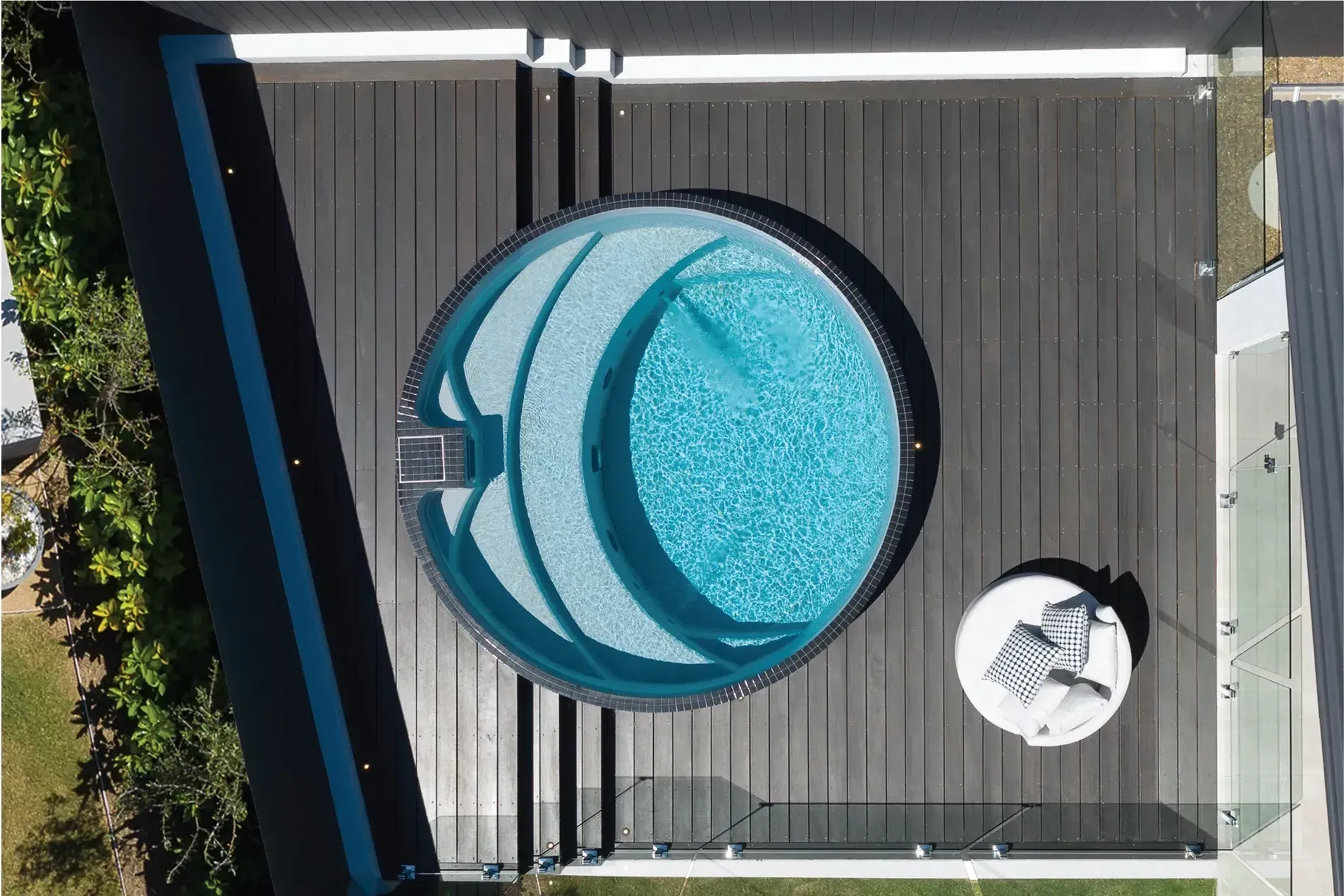 Overhead view of a circular pool with dark wooden decking. A small table with seating is next to the pool.
