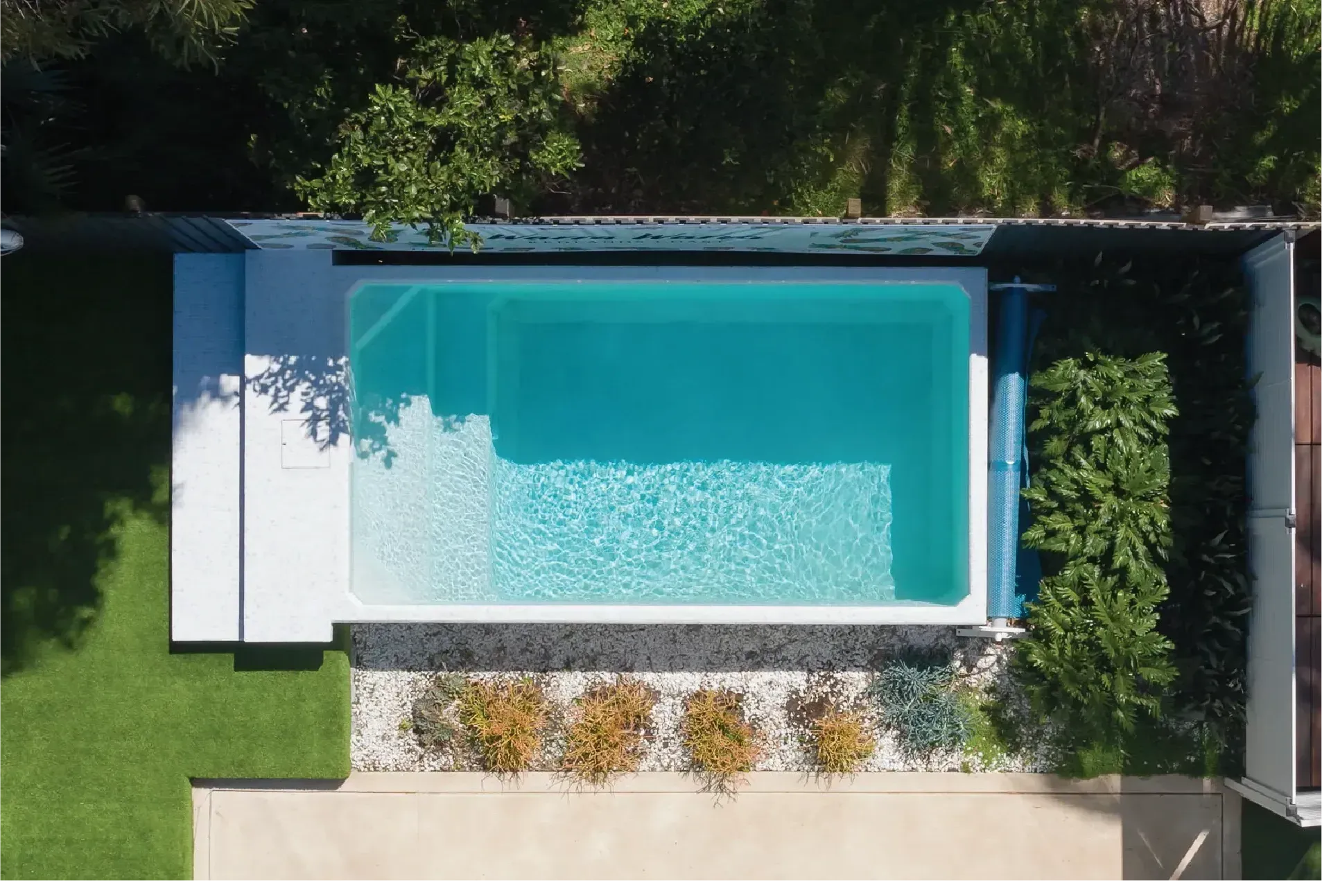 Overhead view of a rectangular pool surrounded by white pavement, green grass, and a small garden bed with colorful plants.