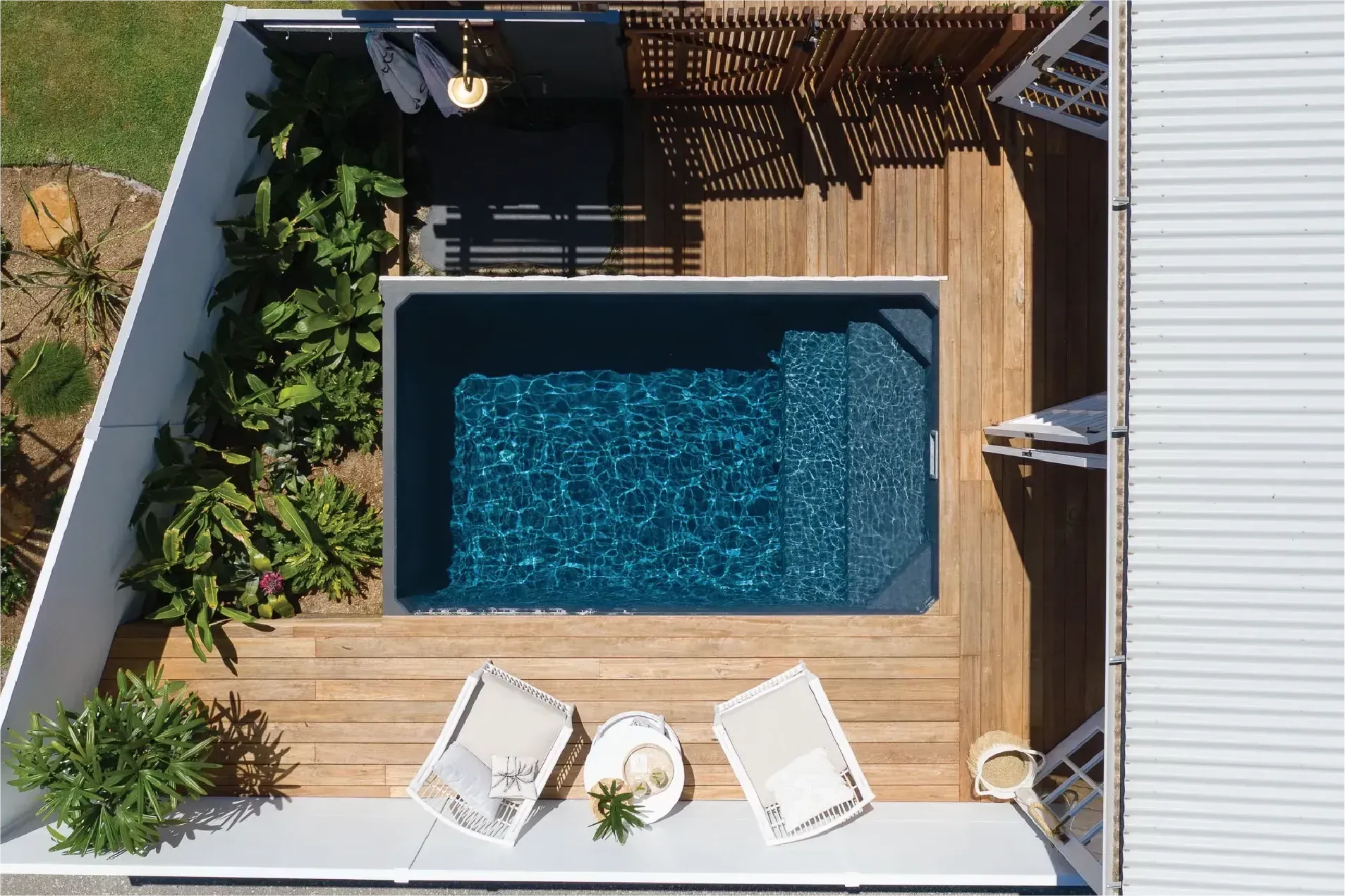 Aerial view of a small backyard pool with a wooden deck, two chairs, and lush landscaping.