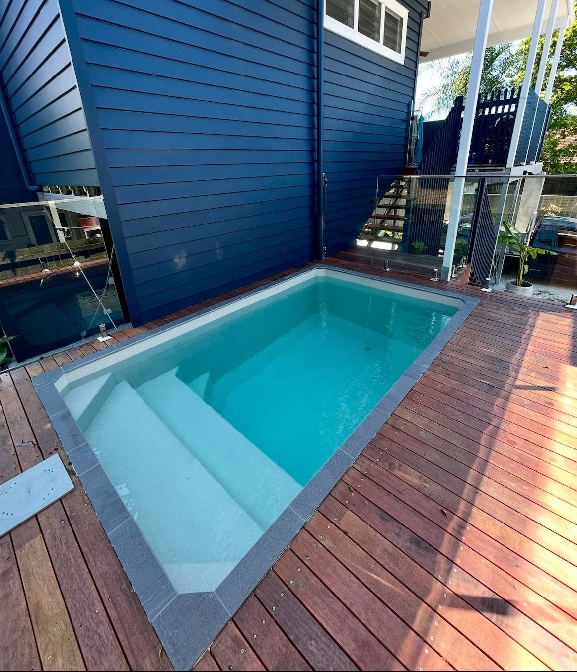 Small, rectangular pool with white steps surrounded by a wooden deck, next to a dark blue house.