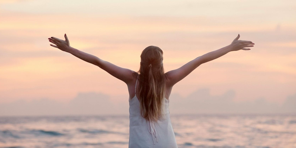 A woman in a white dress is standing on the beach with her arms outstretched.