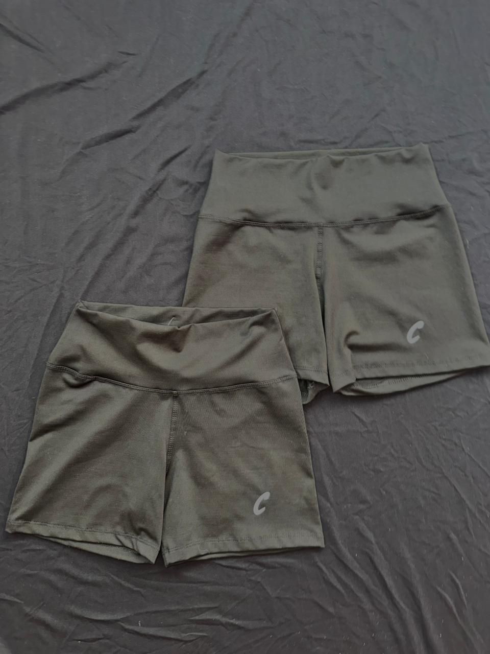 Dos pantalones cortos deportivos verde oliva sobre una sábana gris oscuro. Cada uno luce un pequeño logotipo.
