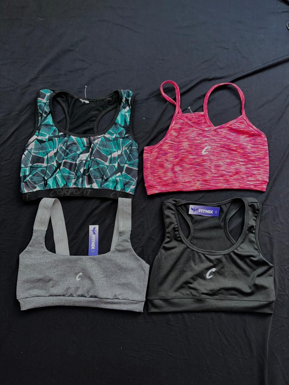 Cuatro sujetadores deportivos en verde azulado, rosa, gris y negro sobre una superficie negra.