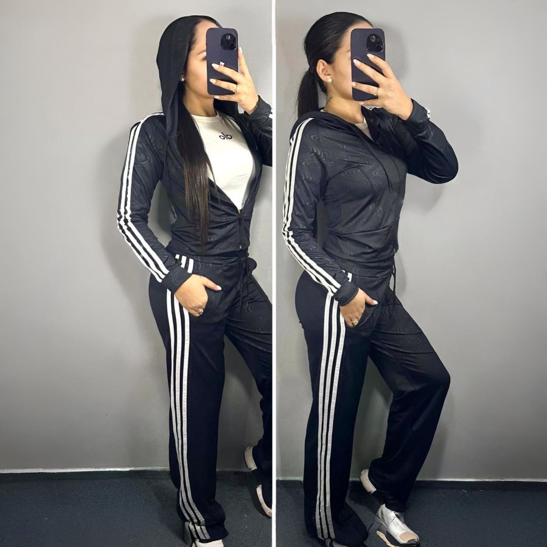 Mujer con chándal Adidas negro, posando frente a una pared gris, sosteniendo un teléfono.