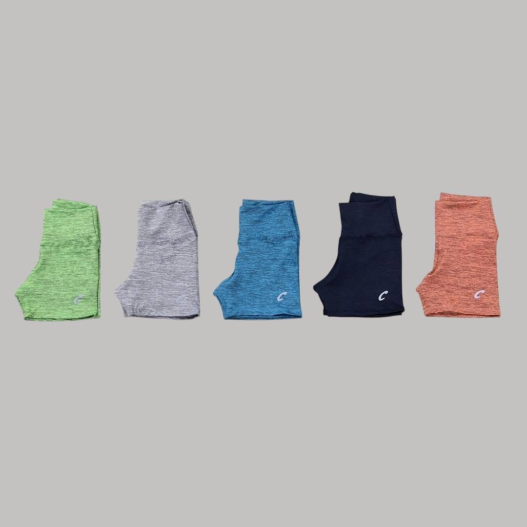 Cinco pares de pantalones cortos de entrenamiento en verde, gris, azul, azul marino y coral, dispuestos sobre un fondo gris.